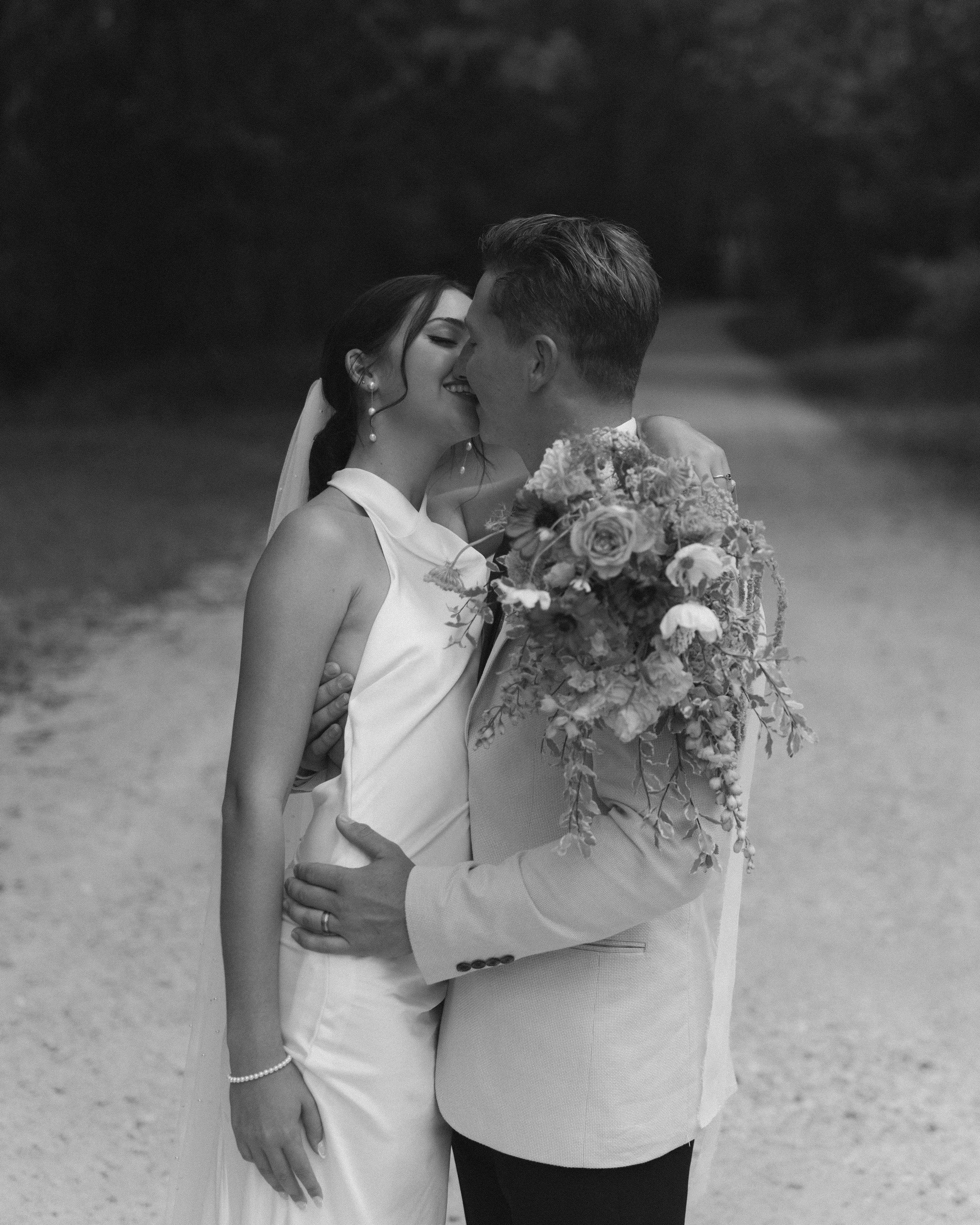A newlywed couple sharing a kiss outdoors, with the bride holding a bouquet of flowers, in a black-and-white photograph.