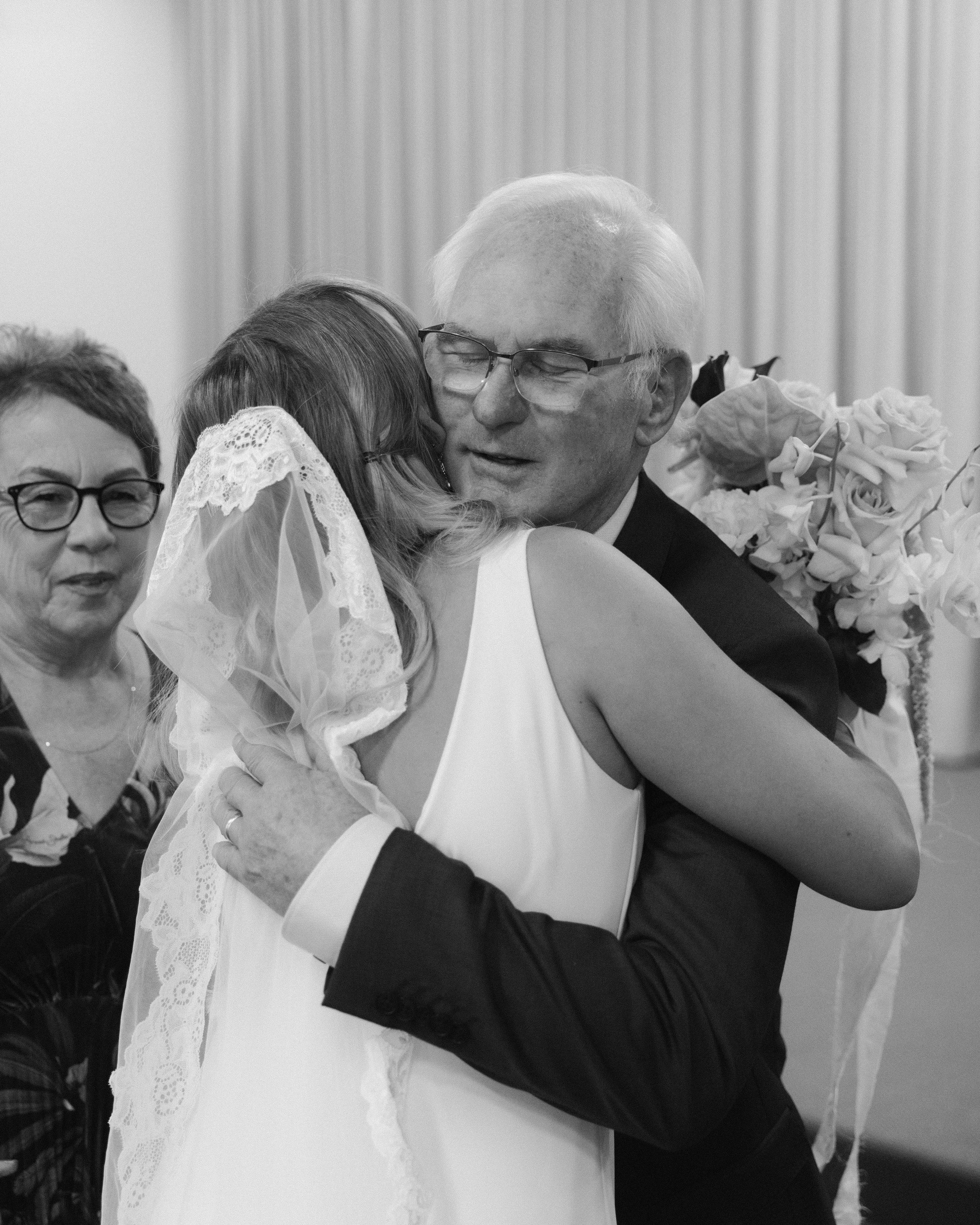 A black and white photo of a wedding scene with a bride hugging an elderly man, who is likely her grandfather, and women observing in the background.