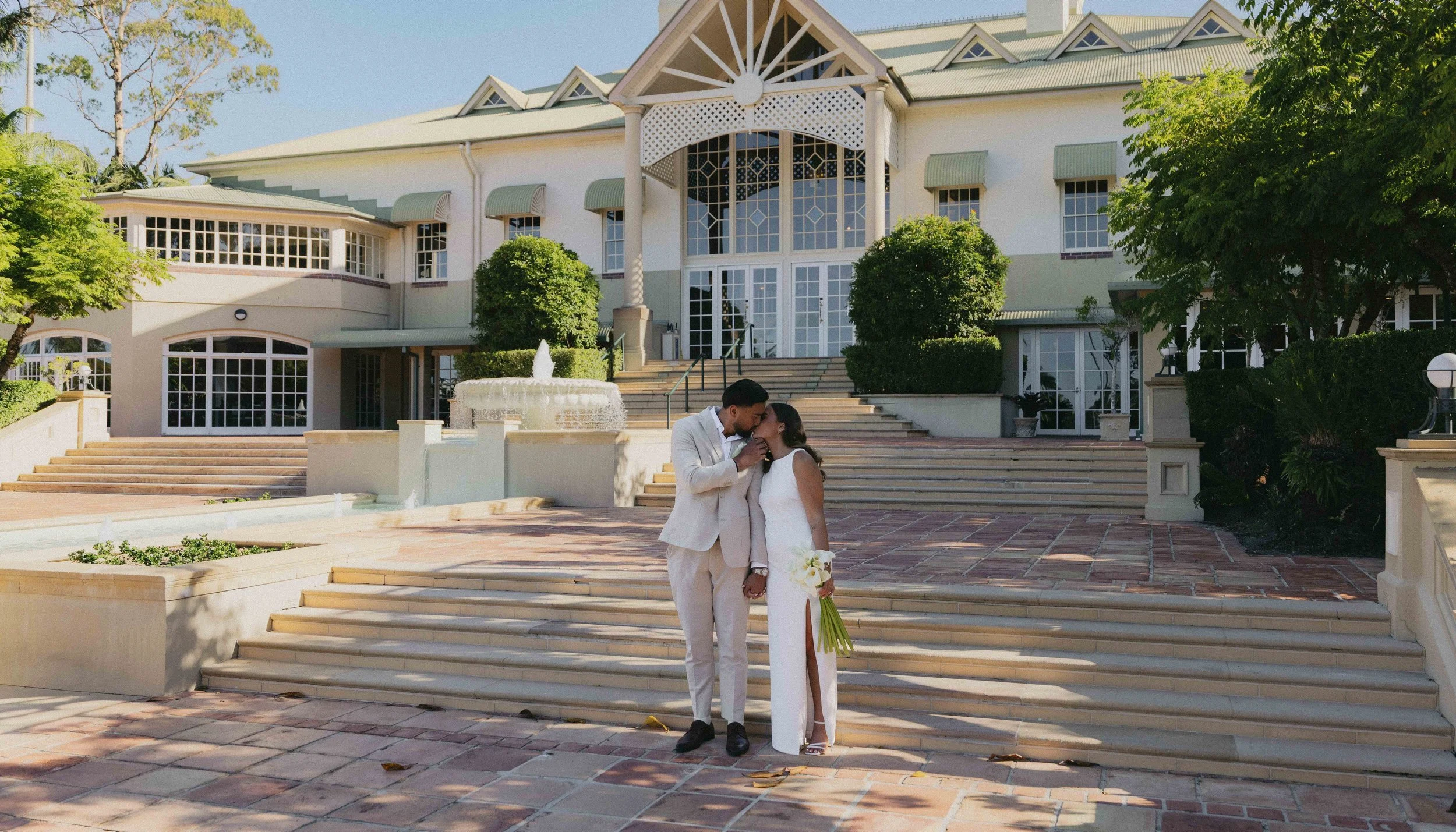 A couple dressed in wedding attire sharing a kiss outdoors in front of a large, white building with a fountain and wide steps.