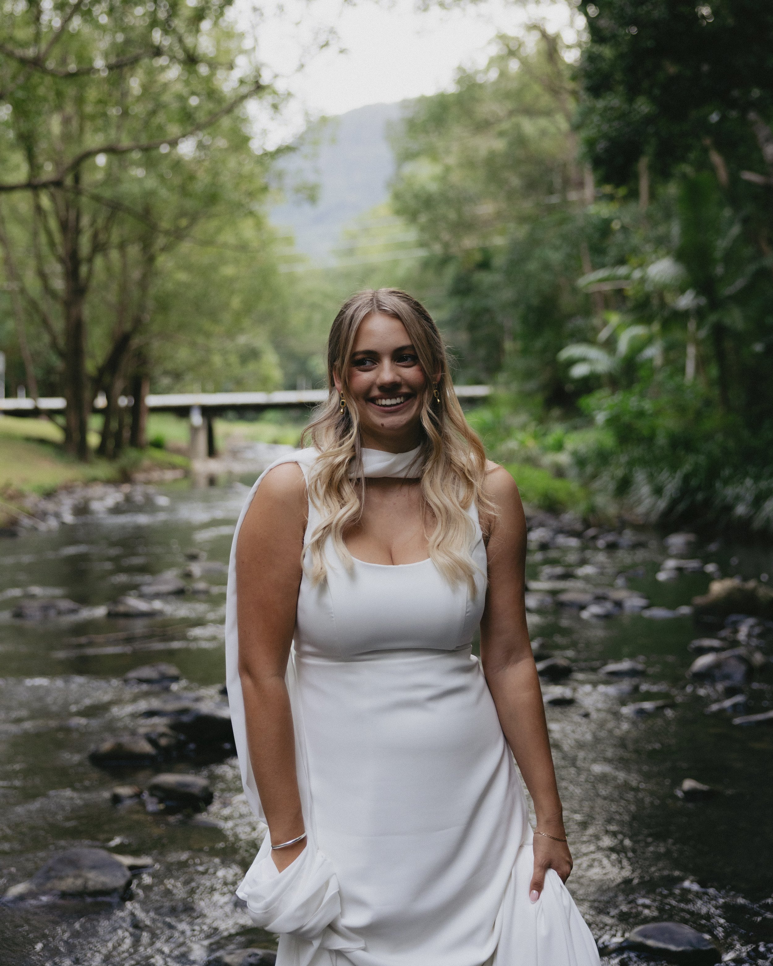 A woman in a white dress standing in a creek with lush green trees and a bridge in the background.