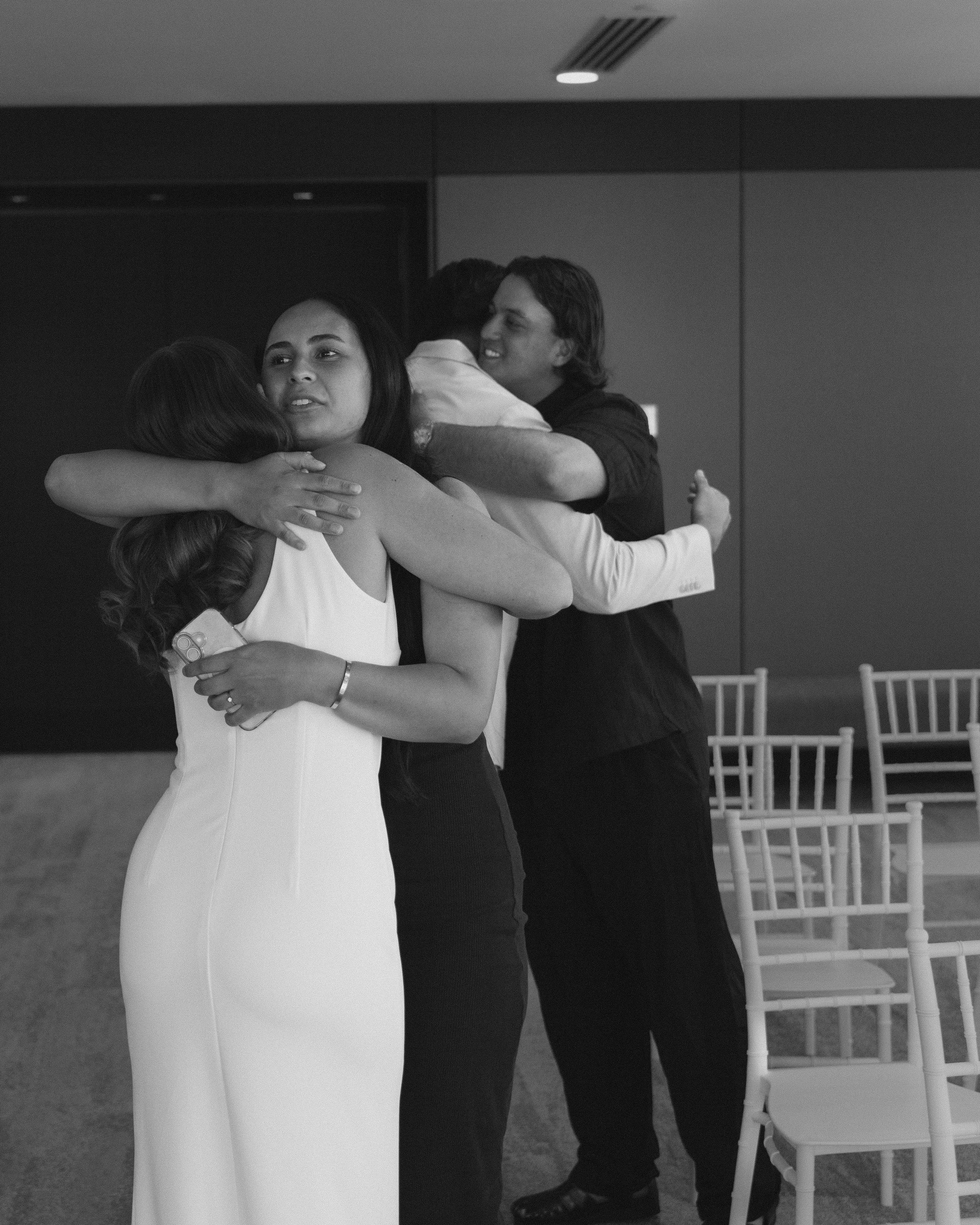 Four people hugging at an indoor event, with empty chairs in the background, black and white photo.