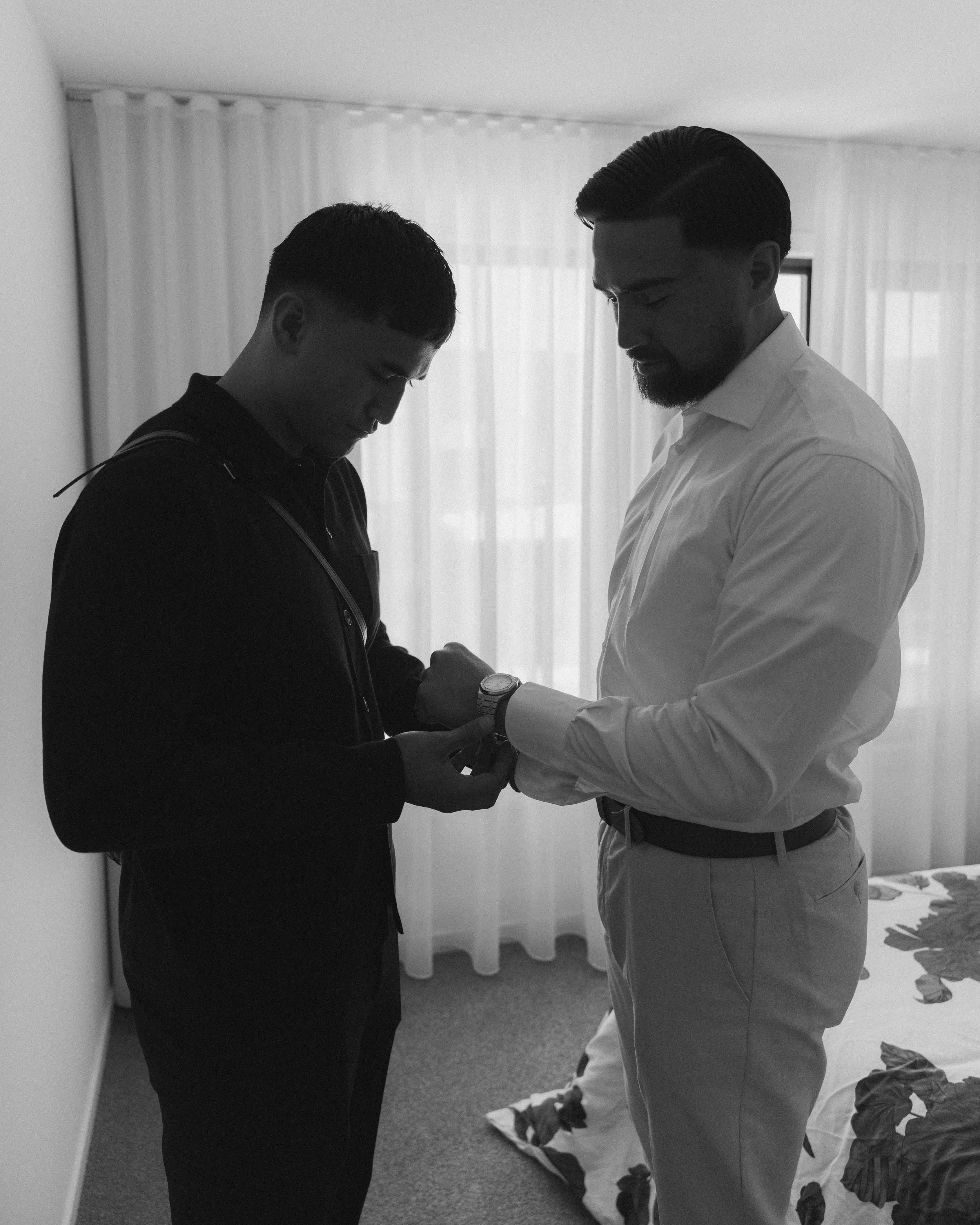 A man in a white shirt helping a young man in a dark jacket with his watch in a hotel room.
