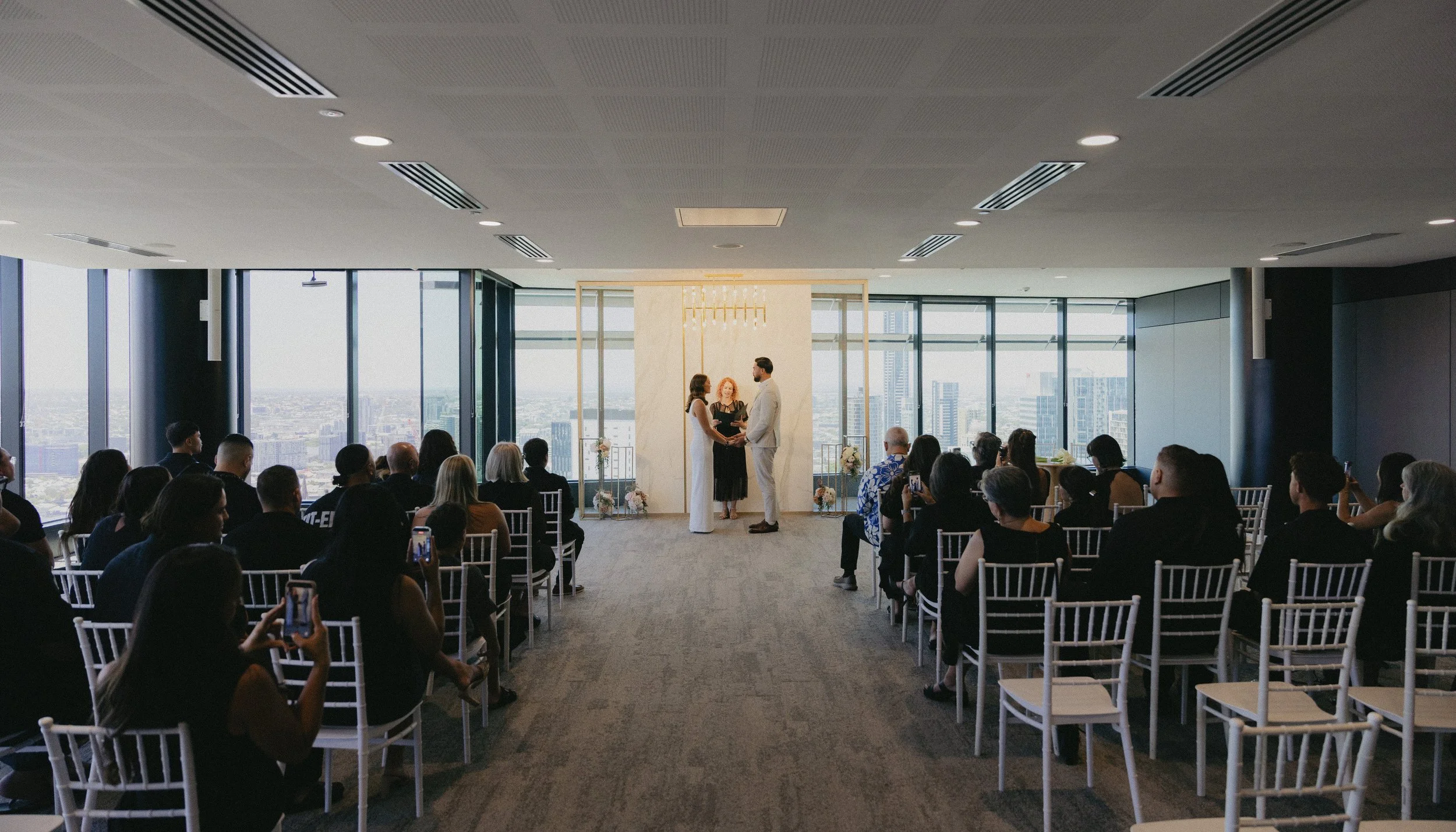 A wedding ceremony taking place in a high-rise building with floor-to-ceiling windows, showing a city skyline, where a couple is standing with an officiant, under a minimalist arch decorated with flowers, with seated guests observing.