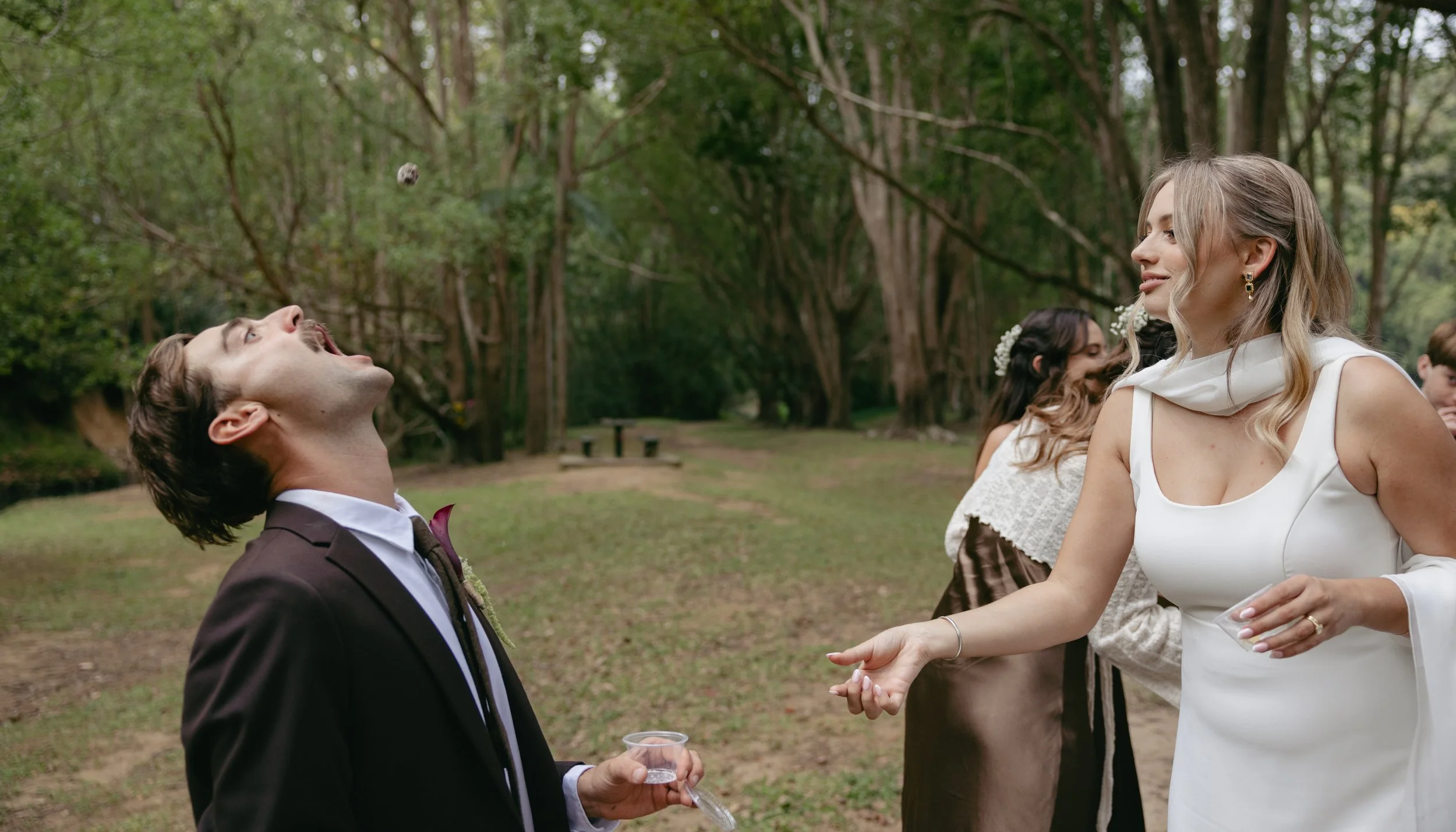 A man in a suit opening his mouth to catch a small object tossed by a woman in a white dress at an outdoor gathering.