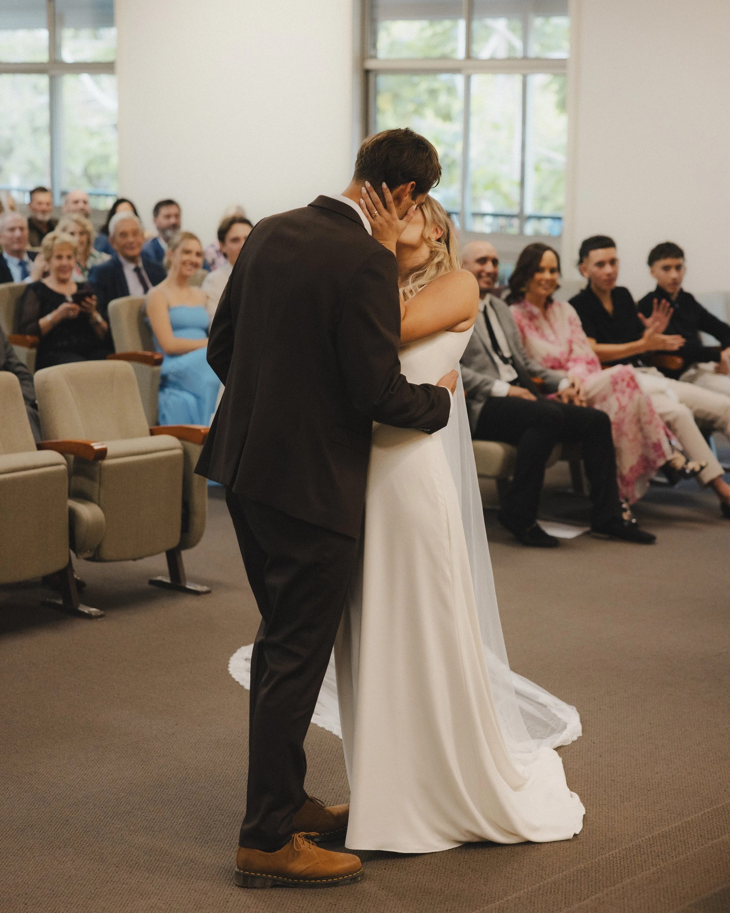 A couple in wedding attire sharing a kiss during a wedding ceremony inside a room with large windows. Guests seated and watching them in the background.
