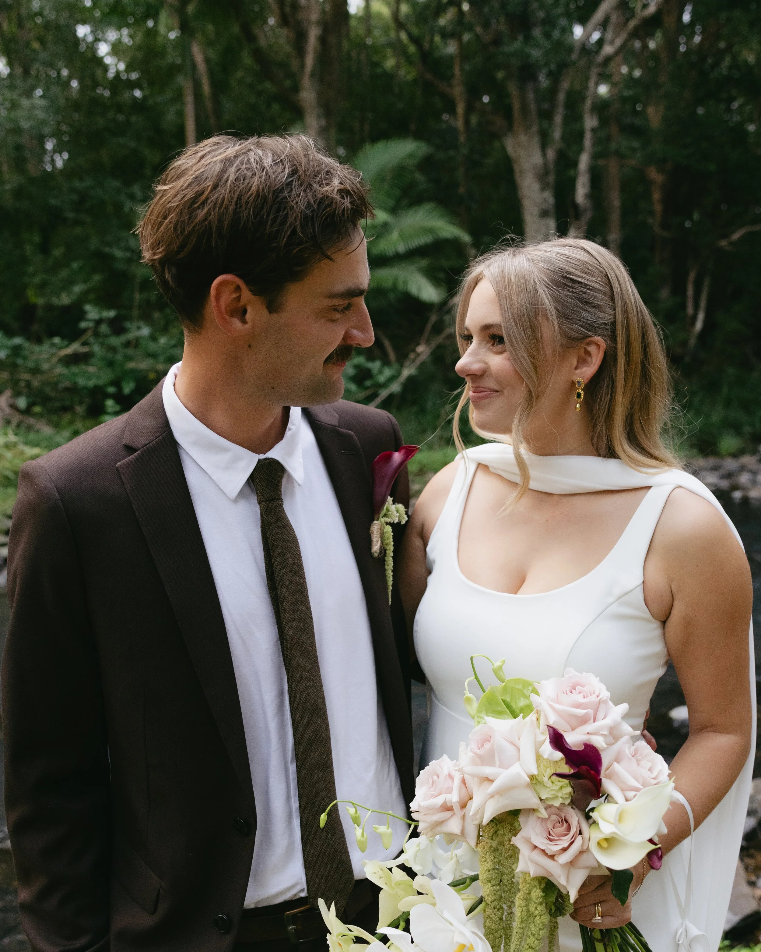 A bride and groom in wedding attire outdoors, smiling and facing each other.