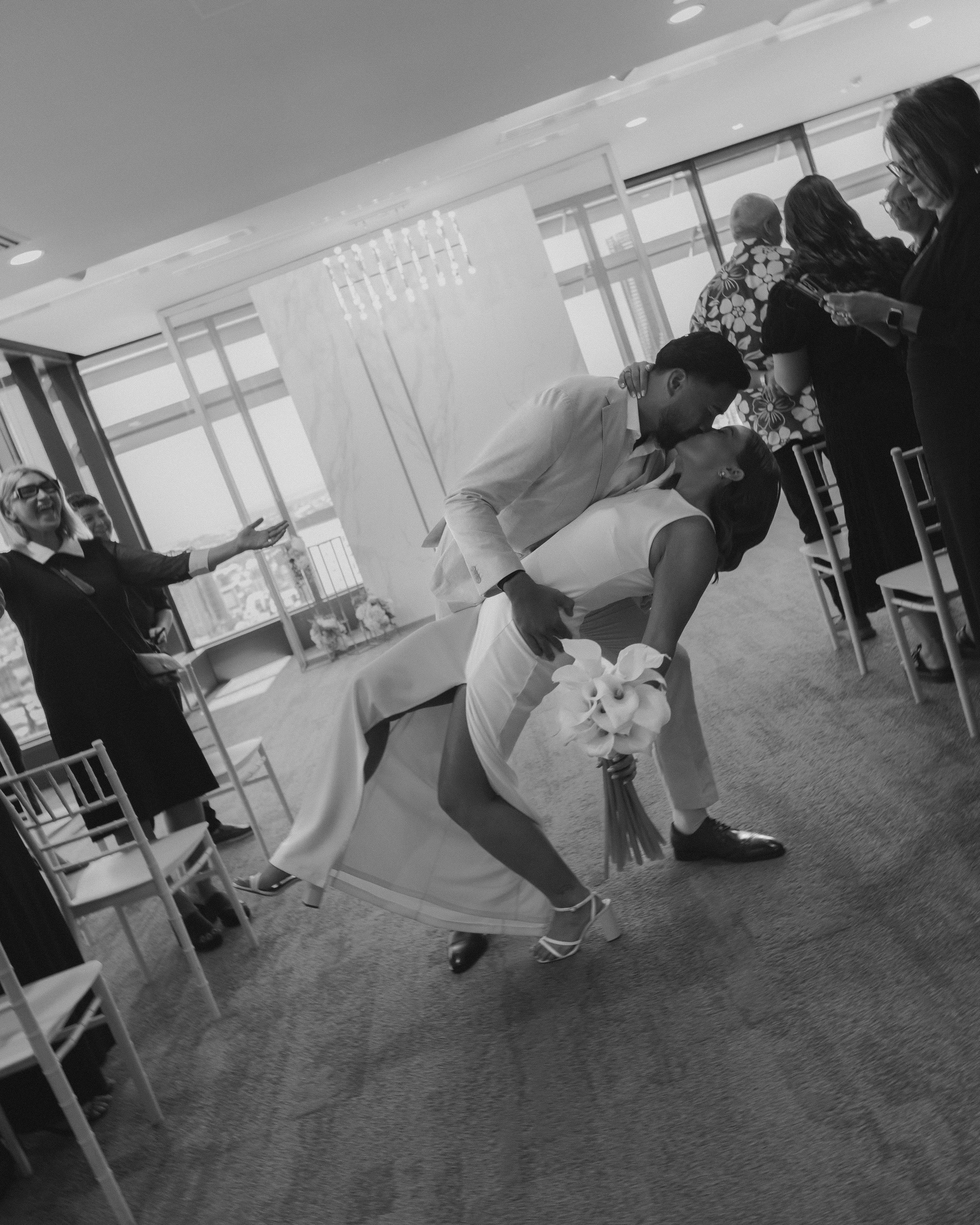 A couple shares a romantic kiss at a wedding reception, with the groom dipping the bride and holding her waist, while the bride holds a bouquet of calla lilies. Guests are seen in the background, some smiling and some looking at their phones, in a bright room with floor-to-ceiling windows.