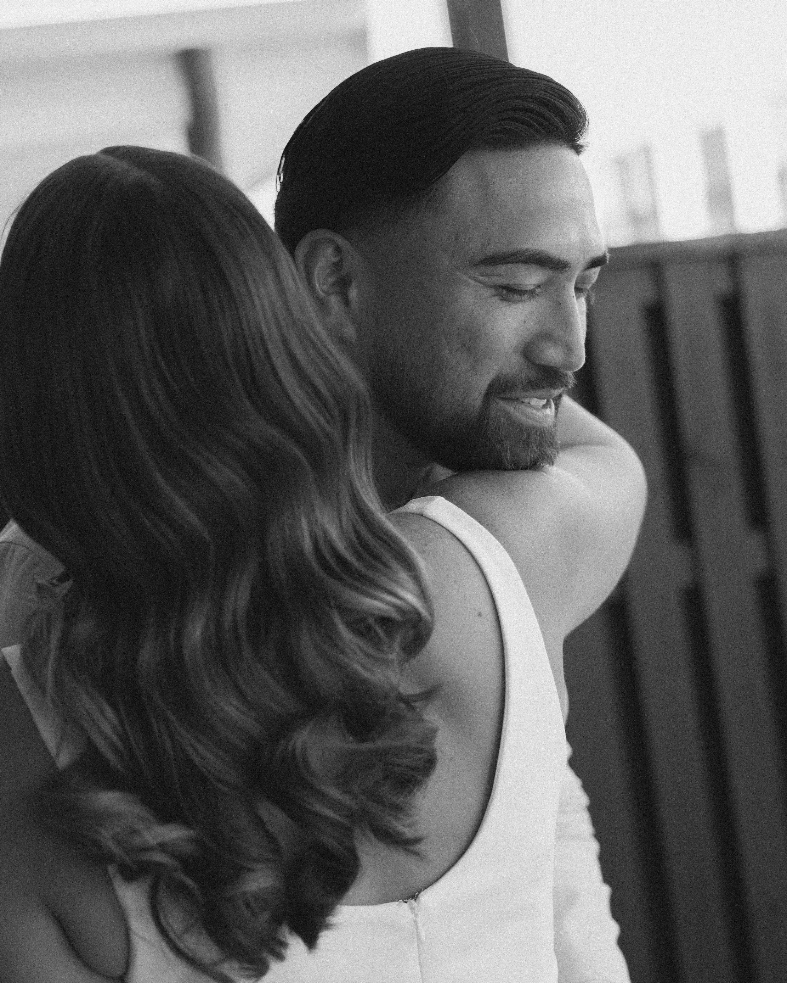 A woman hugging a man with a beard and dark hair, both smiling, in a close embrace.