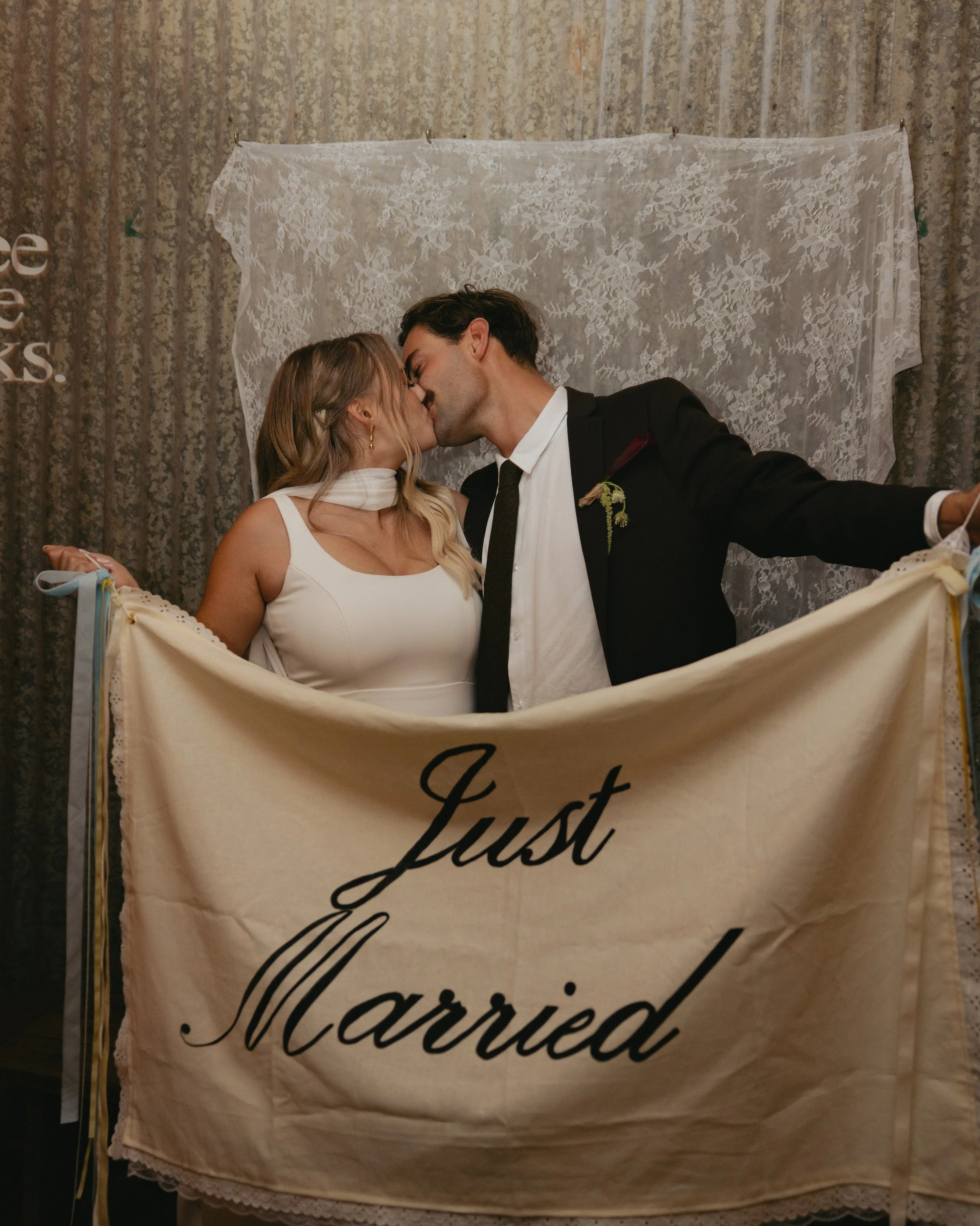 A newly married couple kissing behind a cream-colored "Just Married" banner.