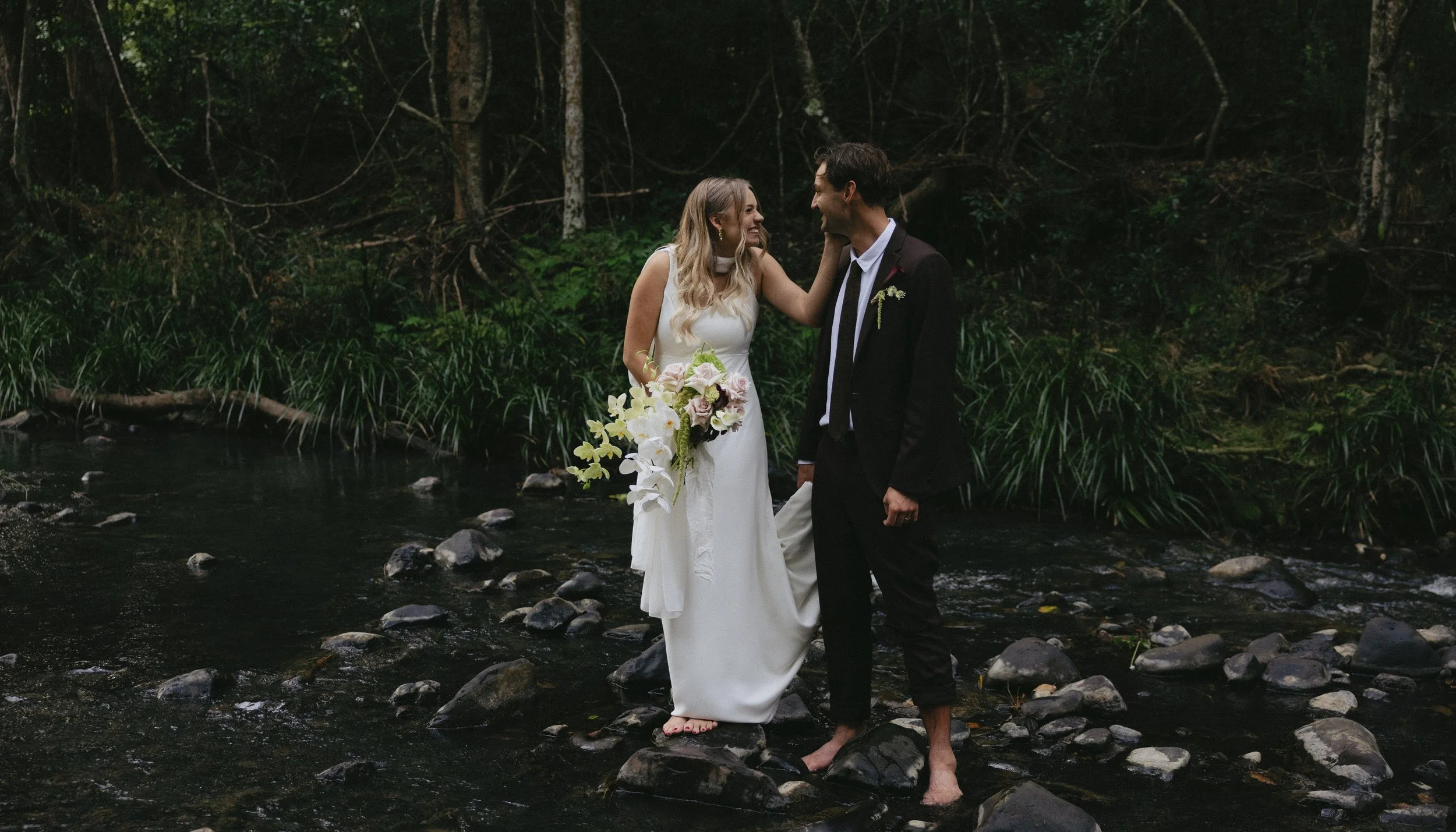 A newlywed couple stands barefoot on rocks in a creek, smiling and looking at each other, with lush green forest in the background.