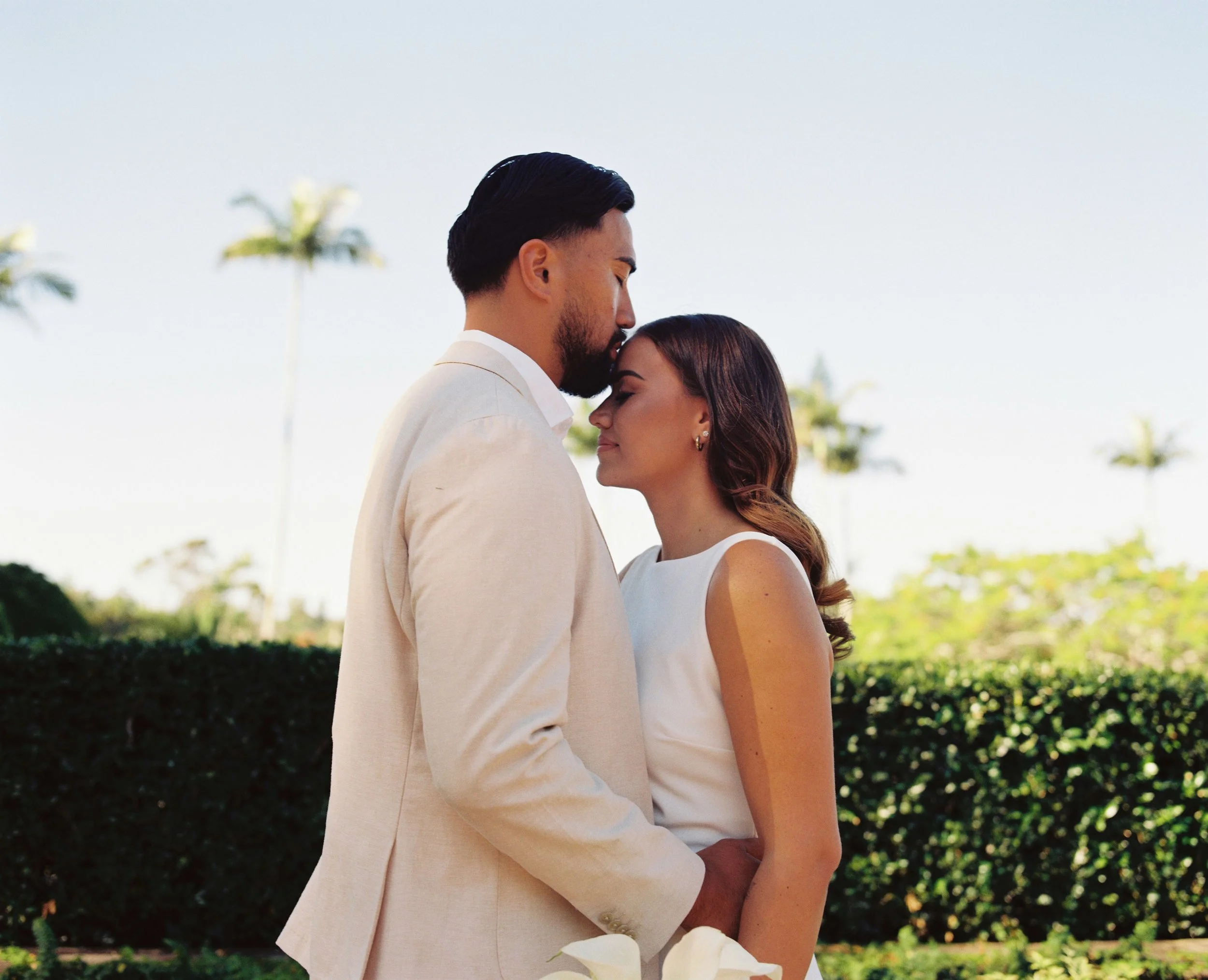 A couple dressed in white, standing close with foreheads touching outdoors, with palm trees and green bushes in the background.