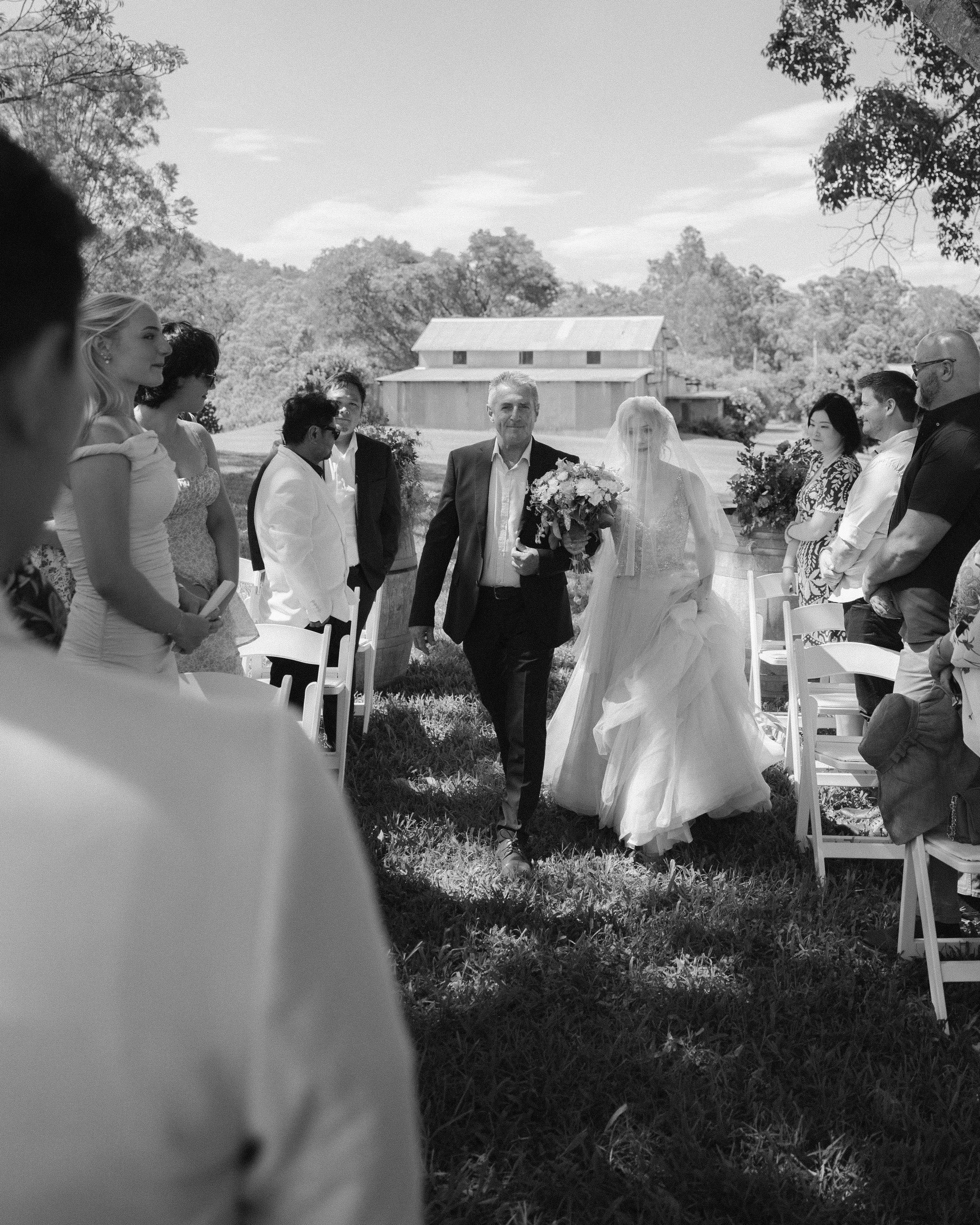 A bride walking down the aisle with her father at an outdoor wedding ceremony, surrounded by guests on a sunny day.