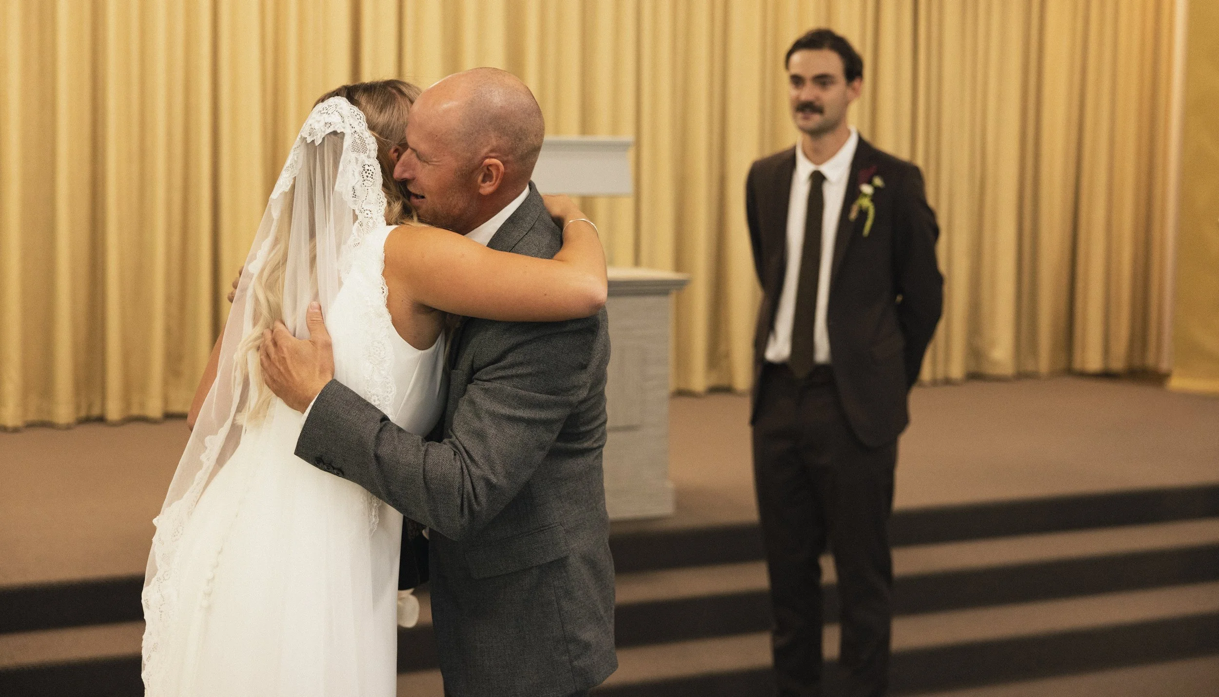 A bride and a bald man in a suit hugging each other during a wedding ceremony, with a man in a suit standing in the background.
