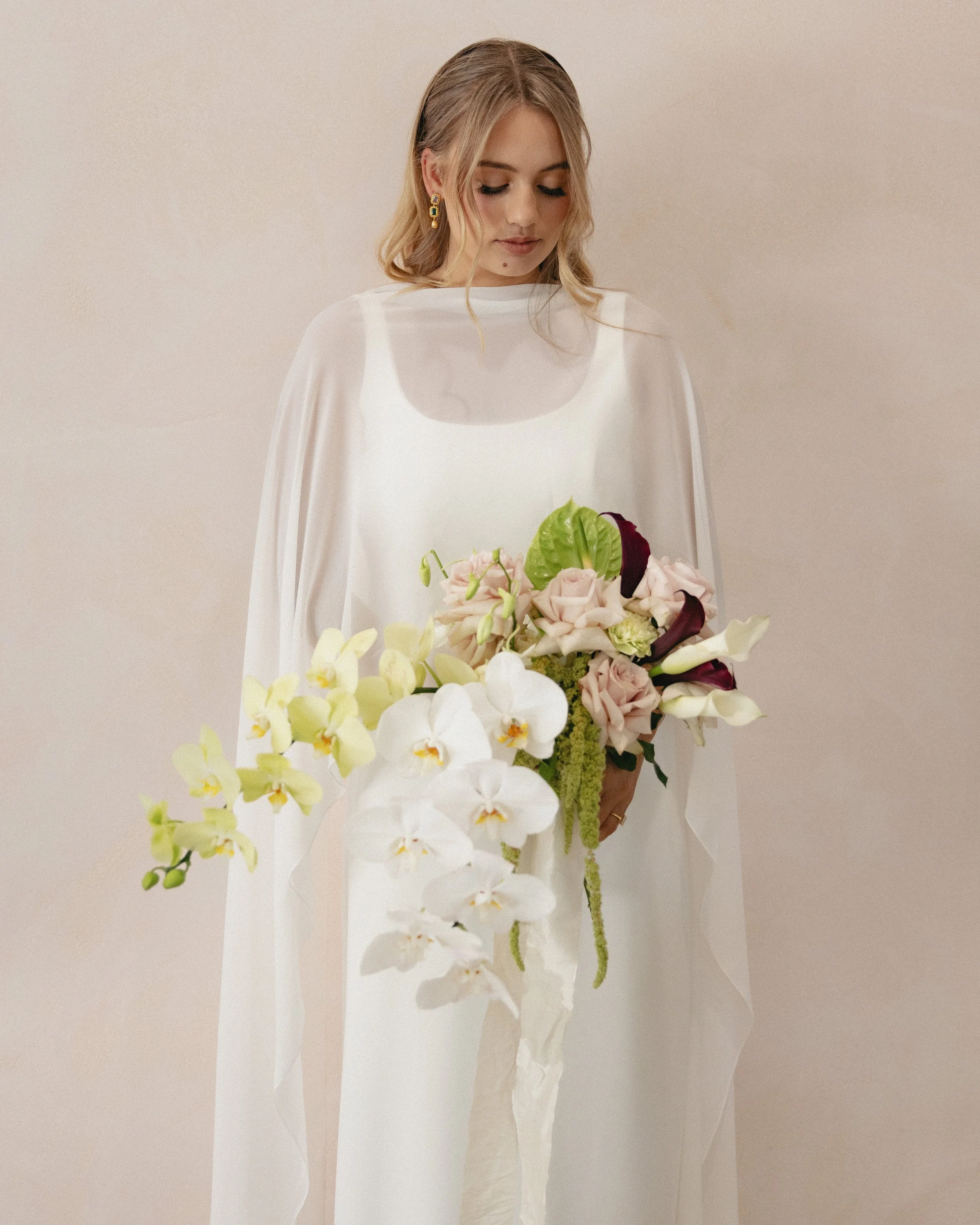 A woman in a white dress holding a large bouquet of pale pink, yellow, purple, and white flowers, including orchids and calla lilies, against a plain beige background.