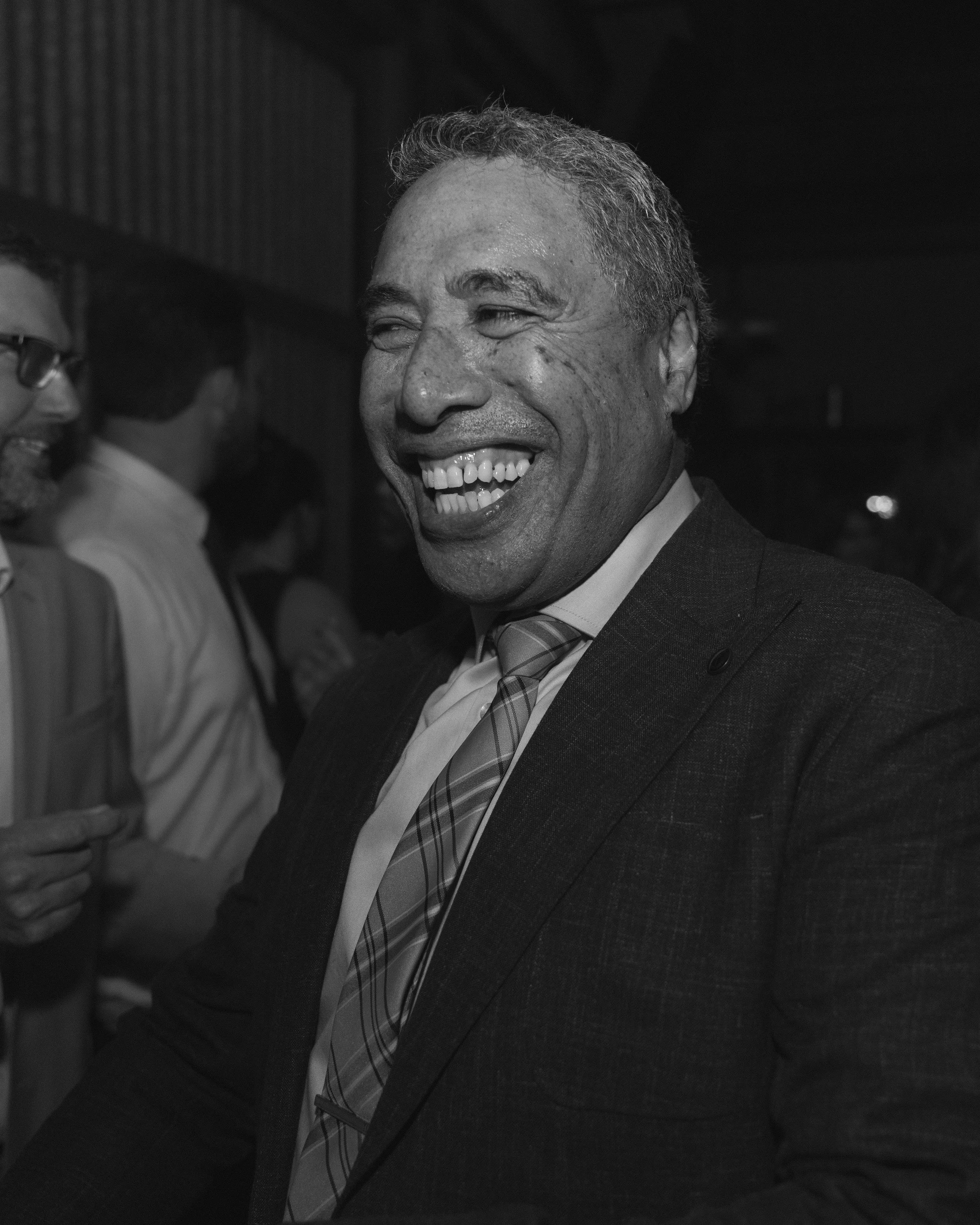 A man with short curly hair and dark skin, dressed in a suit and tie, is smiling broadly at a social event with other people in the background.