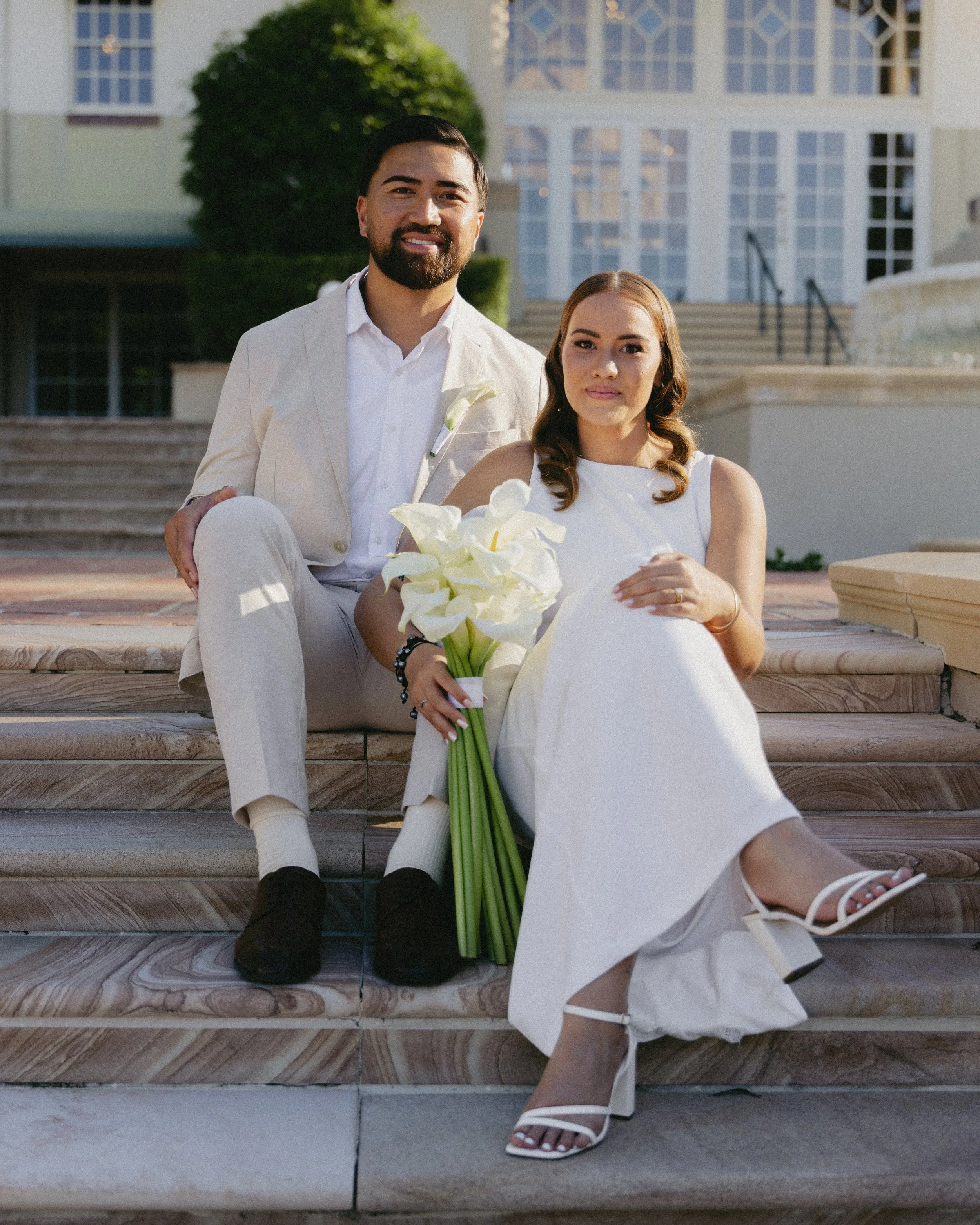 A couple sitting on stairs outside a large house, with the woman holding a bouquet of white calla lilies, both dressed in white, with the woman wearing a white dress and the man in a white suit.