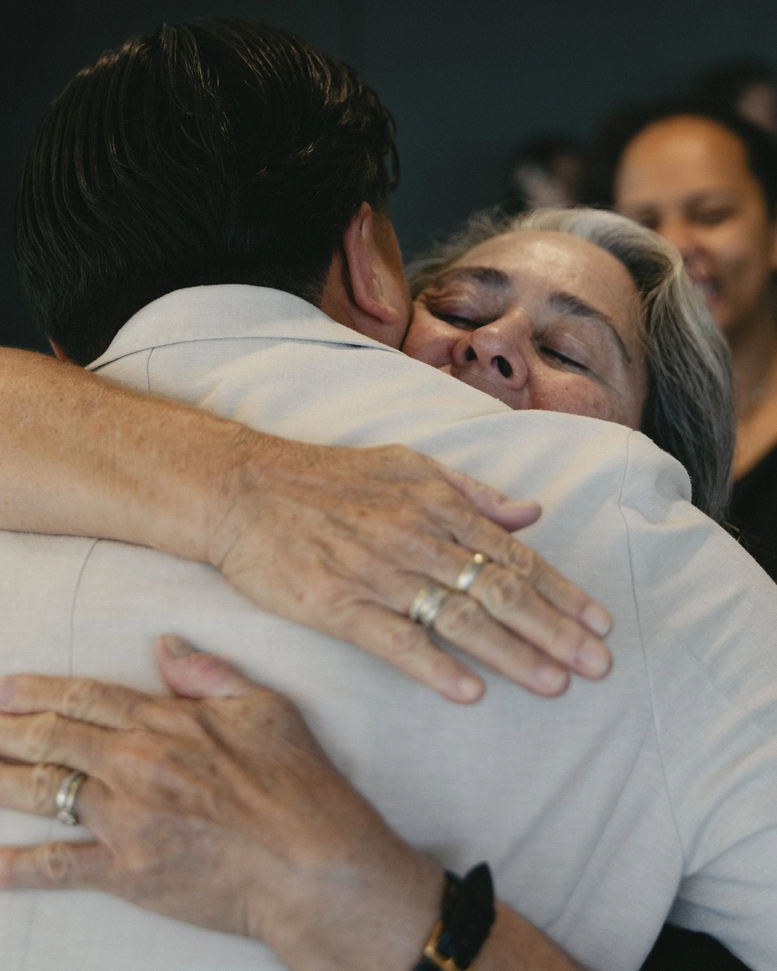 A man and woman embracing in a hug, smiling with eyes closed. The woman has gray hair and is wearing rings, while the man has dark hair and is wearing a white shirt. There are other people in the background.