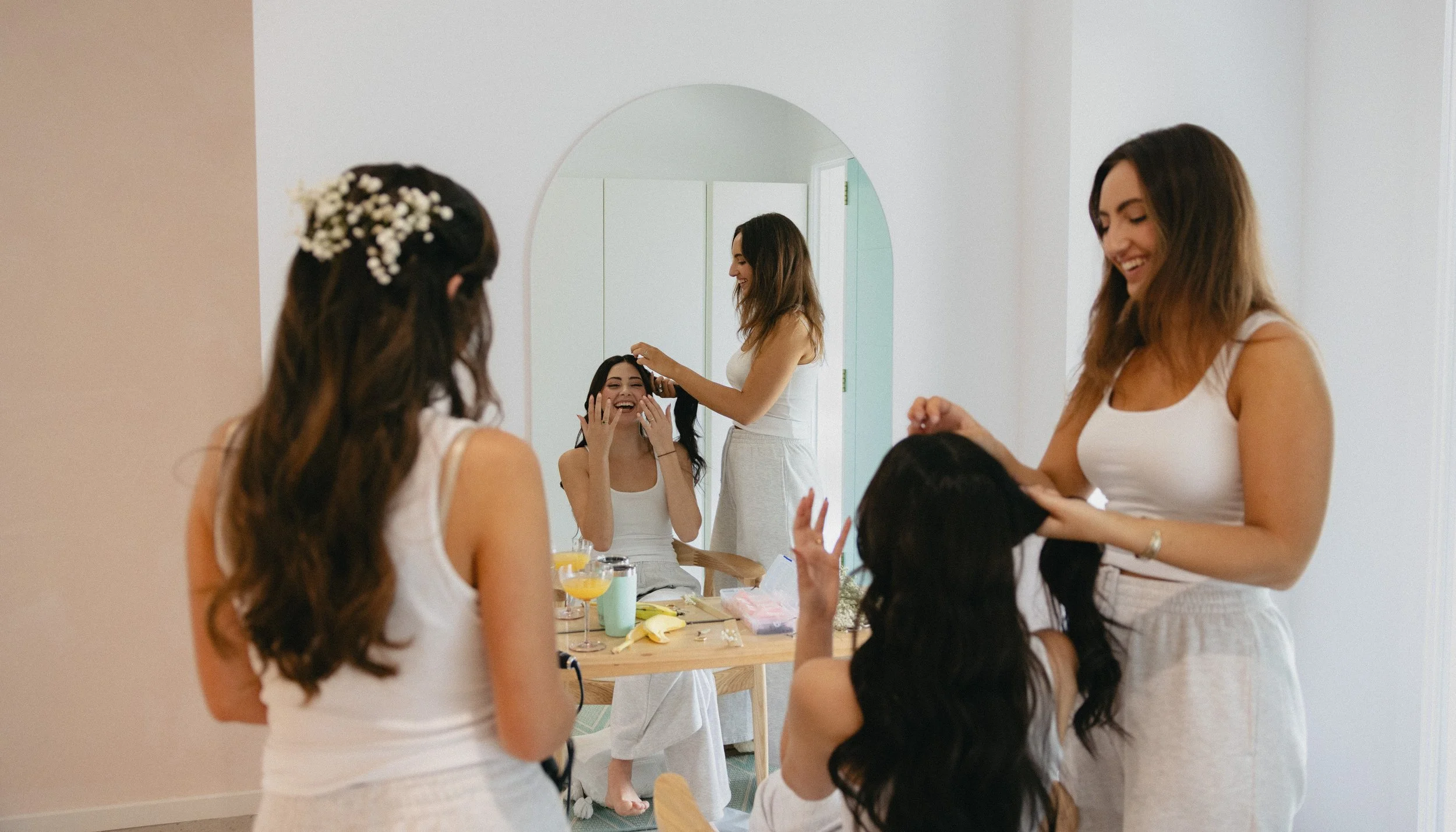 Four women in white outfits engaging in hair and makeup preparations in a bright room with a mirror. One woman is getting her hair done, another is smiling with her hands on her face, and two others are styling hair and smiling.