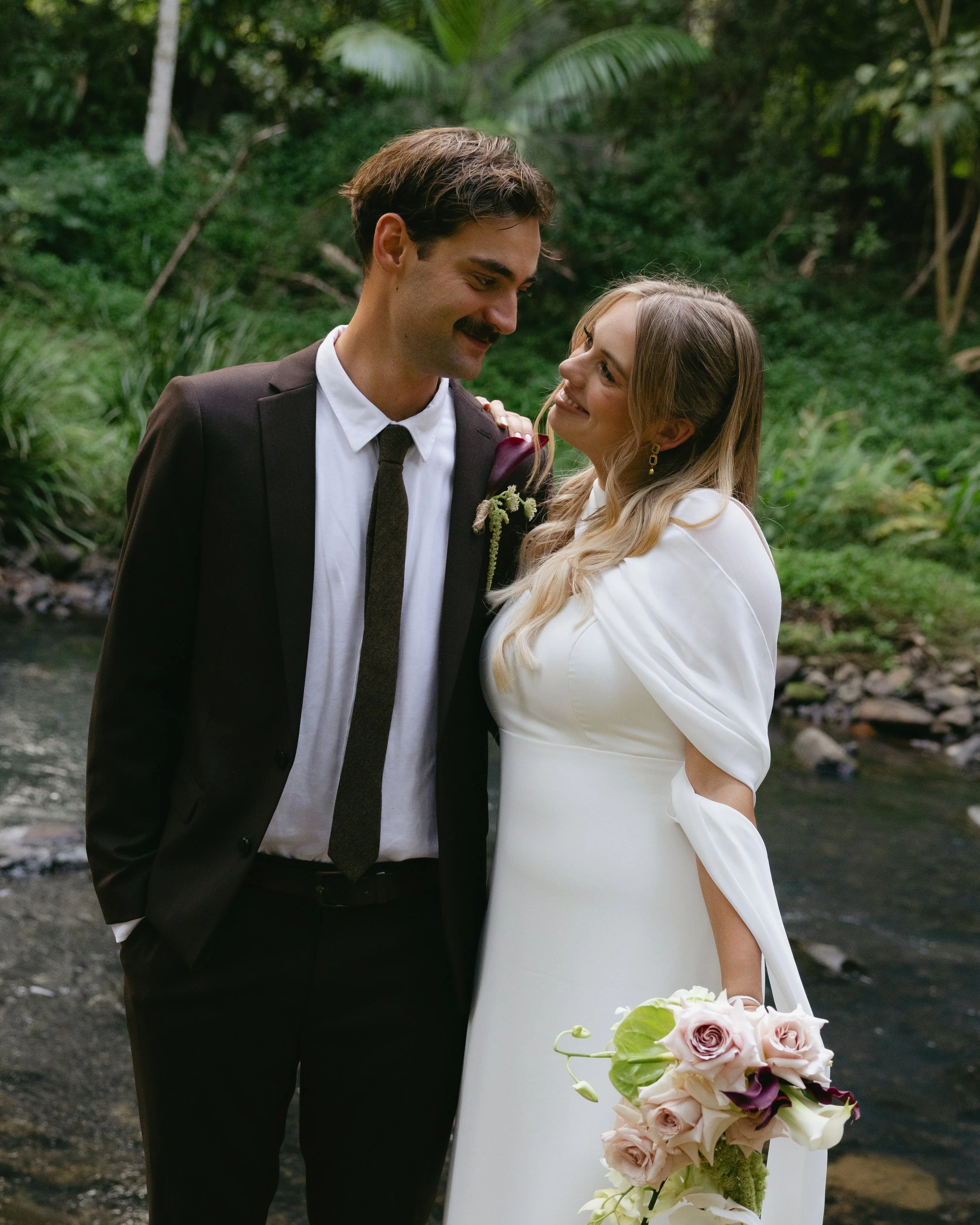 A bride and groom smiling at each other outdoors near a stream in a lush green forest. The bride holds a bouquet of pale pink and dark purple flowers. The groom wears a dark suit with a white shirt and tie, and the bride is in a white wedding dress.