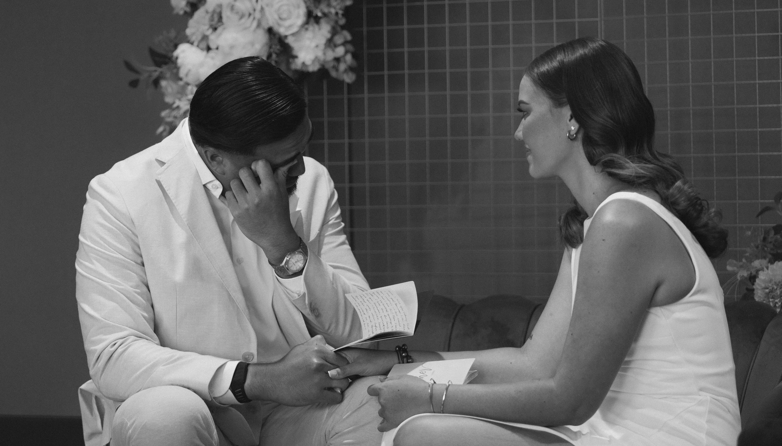 A man and woman seated across from each other, holding hands, in an emotional moment, with the man covering his face with his hand and the woman smiling gently, in a room with floral decorations in the background.