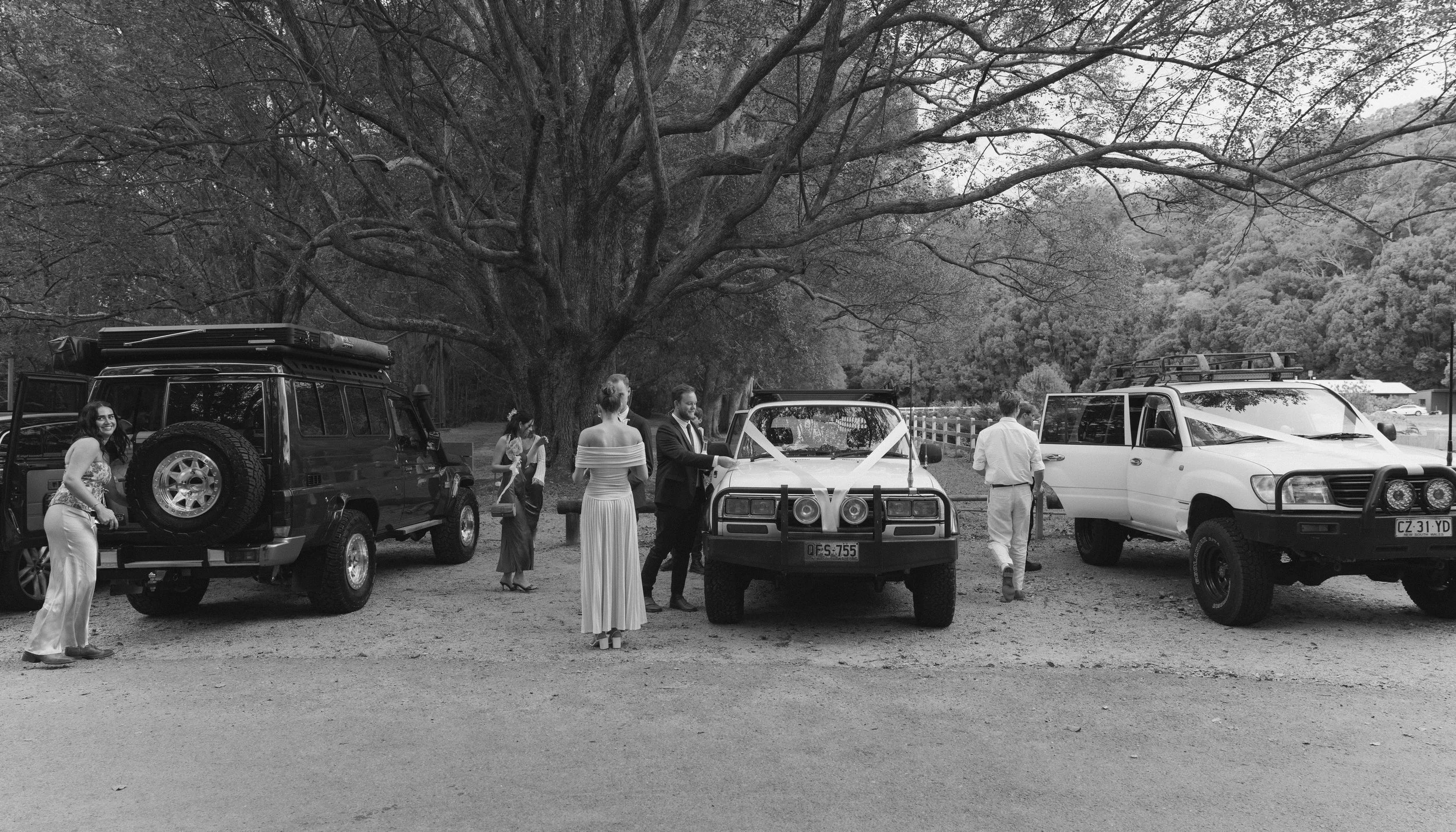 Black and white photo of people gathering outdoors near old vehicles and a large tree, with some people dressed semi-formally.