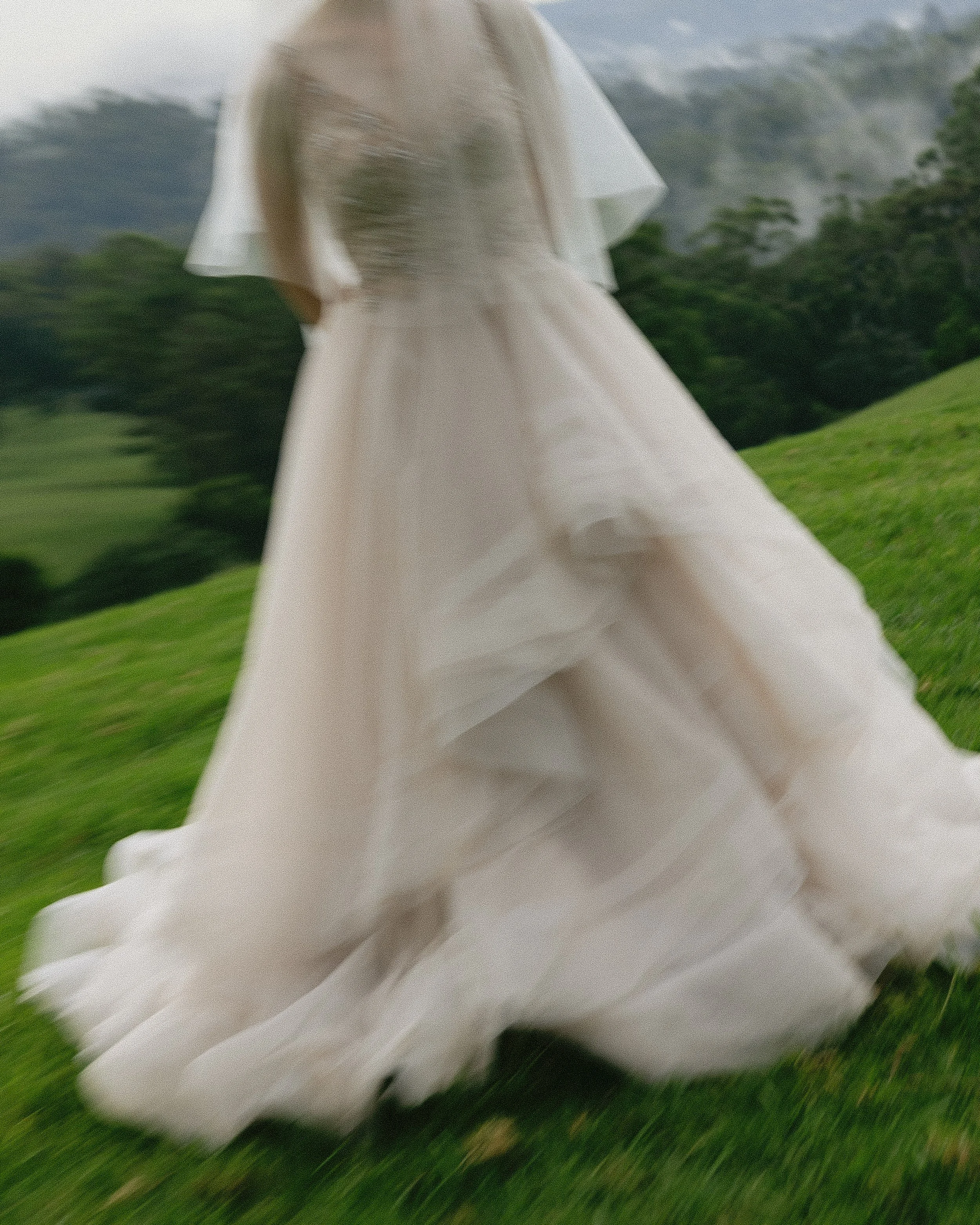 A person in a flowing white dress walking through a grassy green field with mountains in the background.