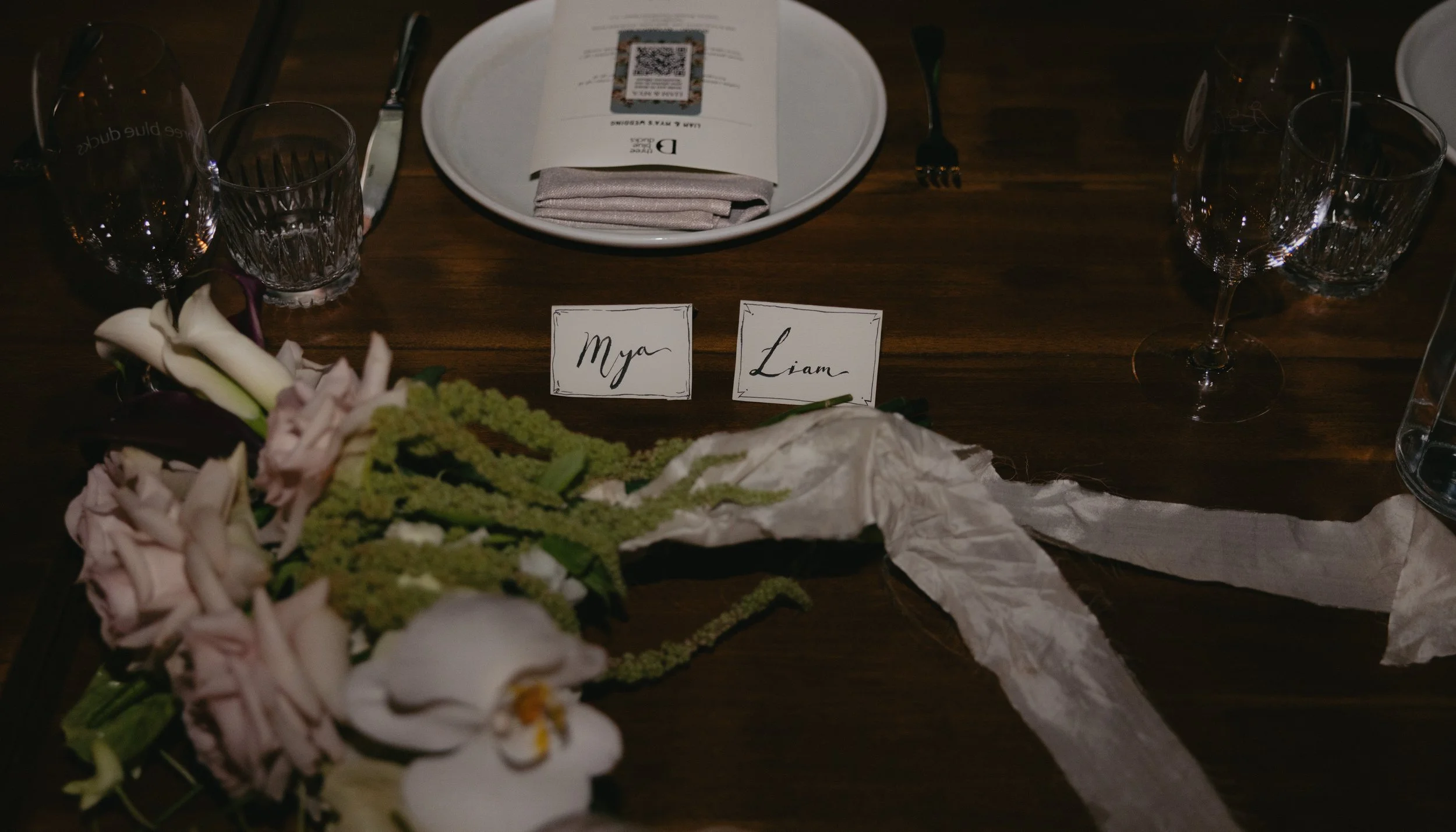 A wedding reception table setting with a floral arrangement, place cards reading "Mya" and "Liam," wine glasses, a plate with a folded napkin, and a menu with a QR code on top.
