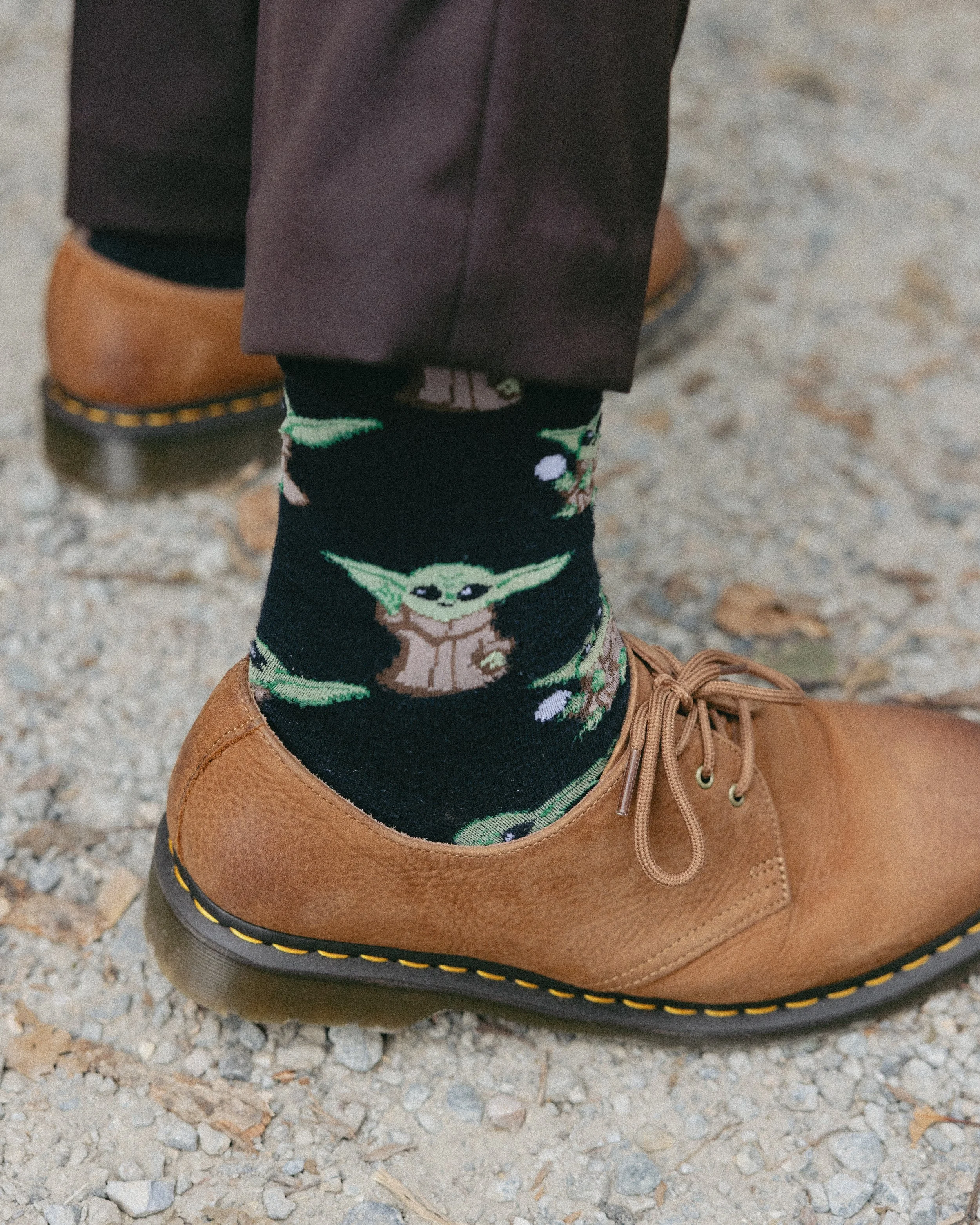 Person wearing brown shoes, black pants, and black socks with Baby Yoda designs, standing on gravel ground.