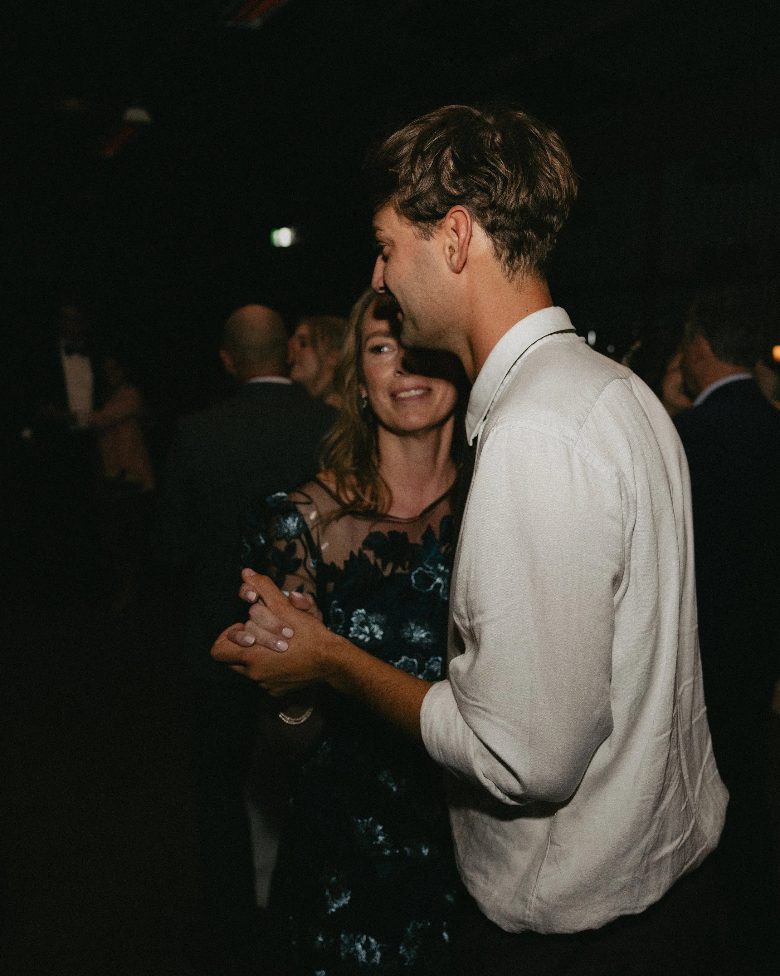 A man and woman dancing together at a social event, with other people visible in the background.