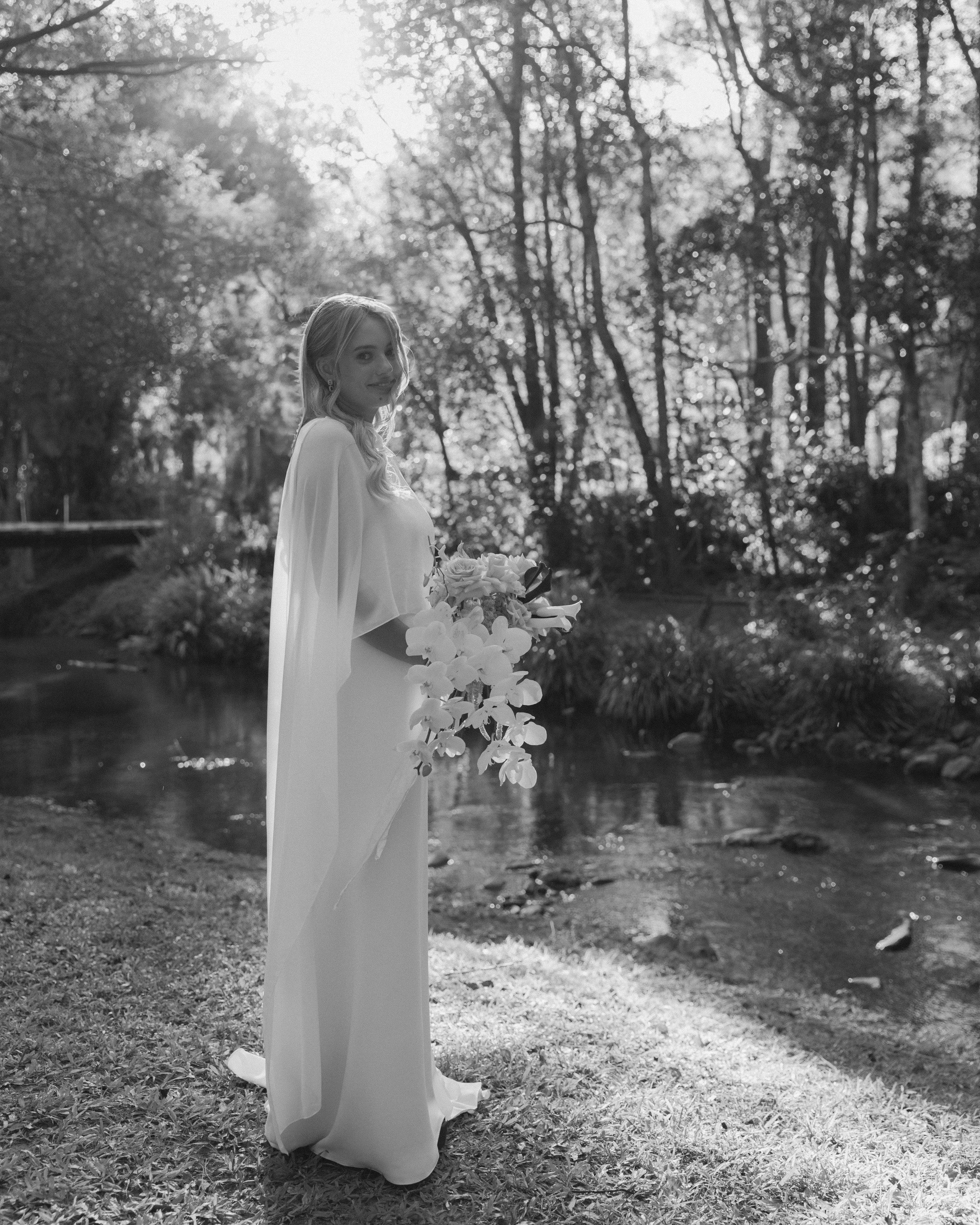 A woman in a white wedding dress holding a bouquet of flowers, standing outdoors near a stream with trees in the background, black and white image.