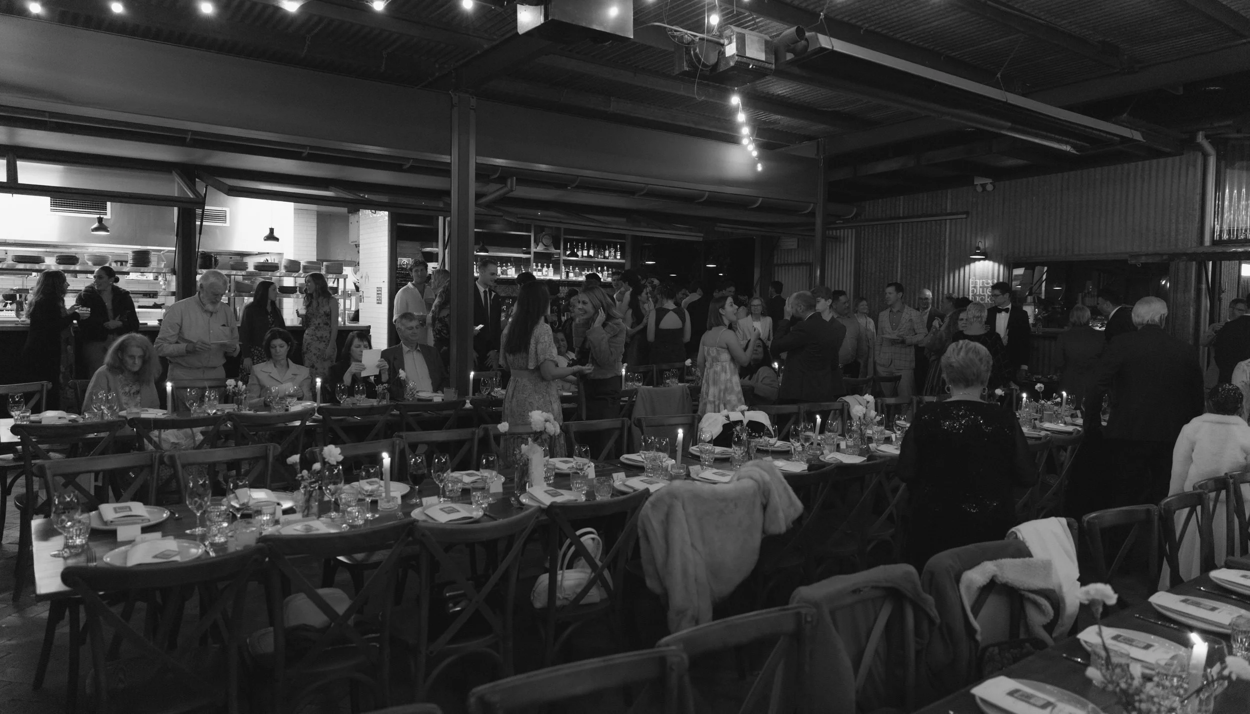 People gathered in a restaurant or banquet hall, some standing and talking, others sitting at decorated tables with plates, glasses, and flowers, in a black-and-white photo.