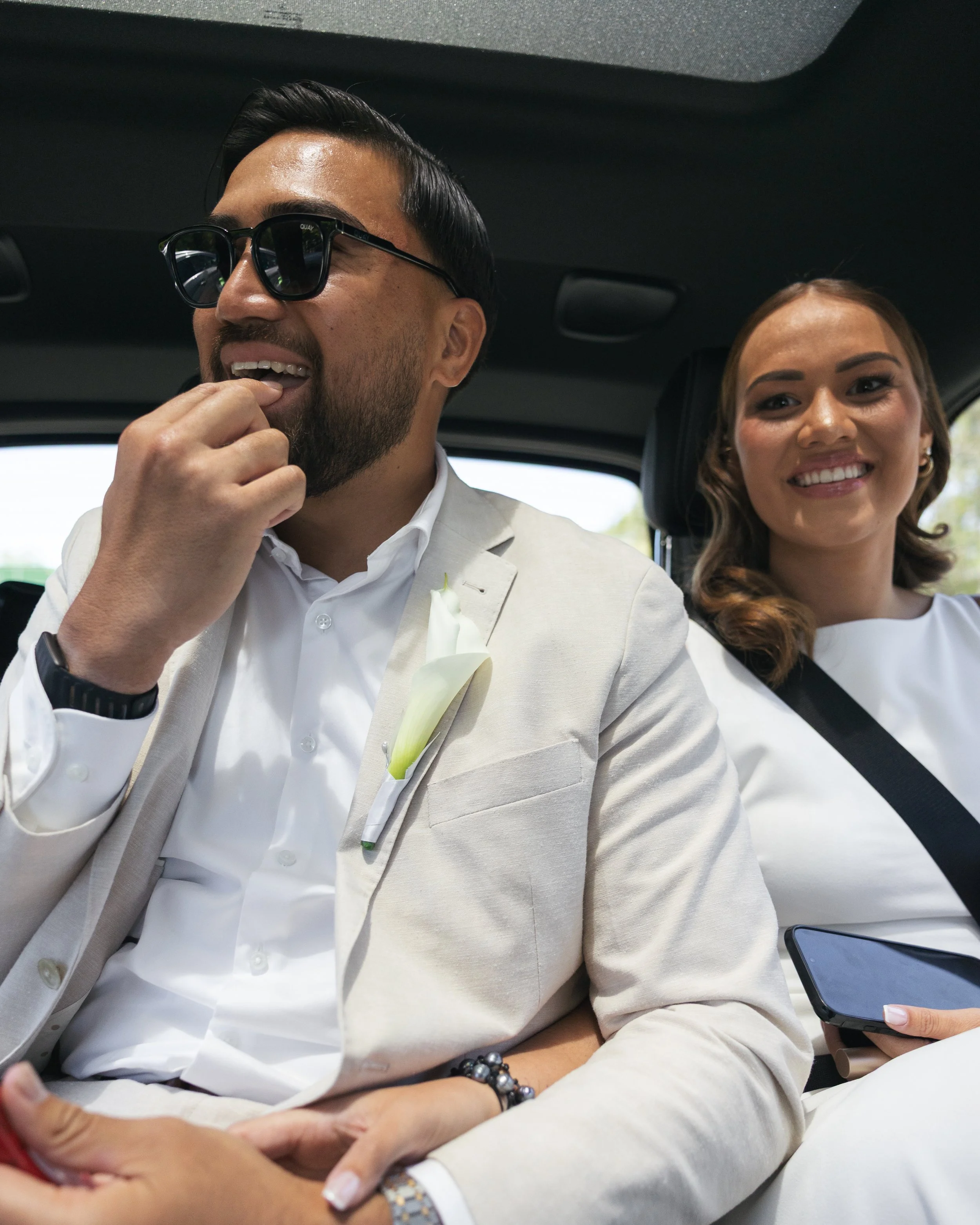 Two people seated inside a vehicle, dressed in attire suitable for a wedding or special event. The man is wearing sunglasses and a light-colored suit with a white shirt, and has a boutonniere on his lapel. The woman behind him is smiling, wearing a white dress, and holding a smartphone.