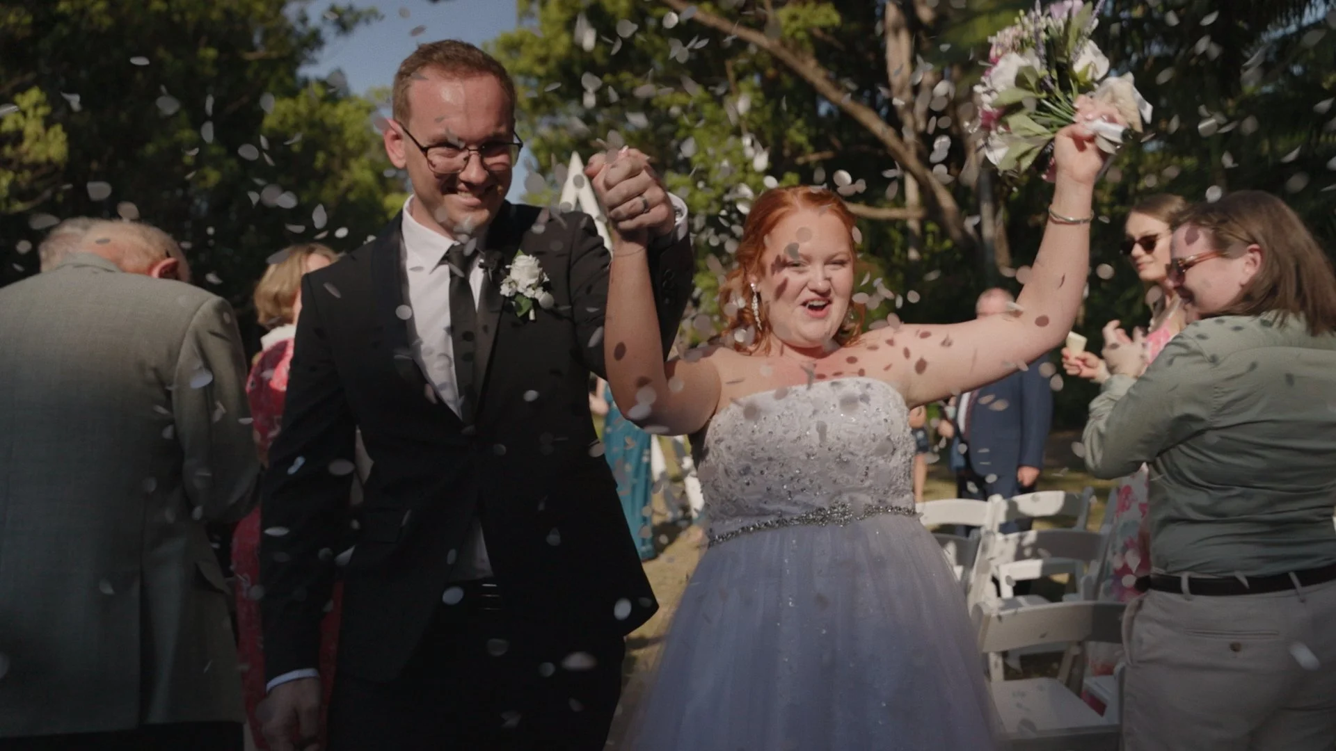 A bride and groom celebrating at their wedding, holding hands and raising a bouquet, with guests behind them and confetti in the air.