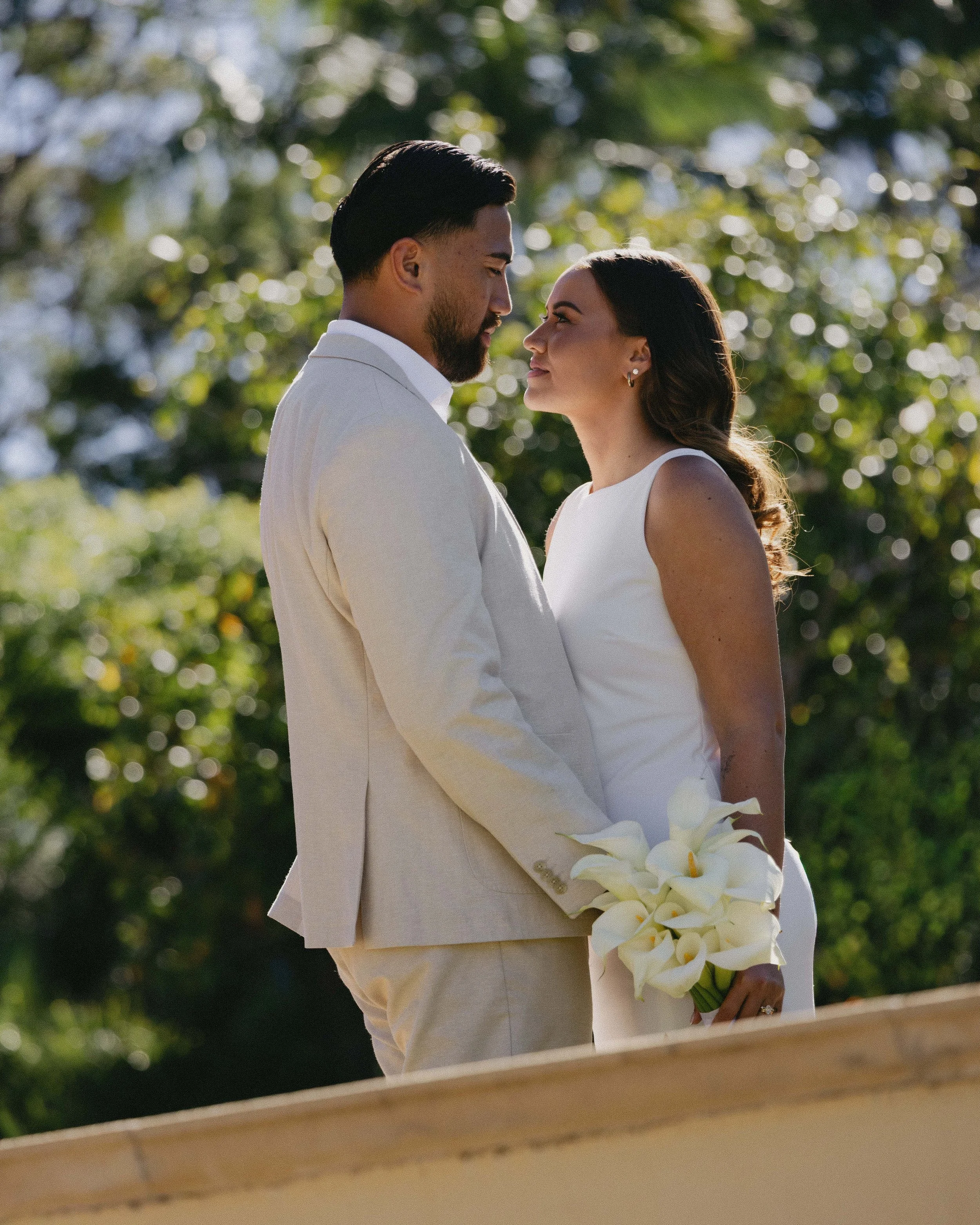 A newly married couple standing close together outdoors, with the groom in a light-colored suit and the bride in a white dress holding a bouquet of white calla lilies. They are gazing into each other's eyes with trees in the background.