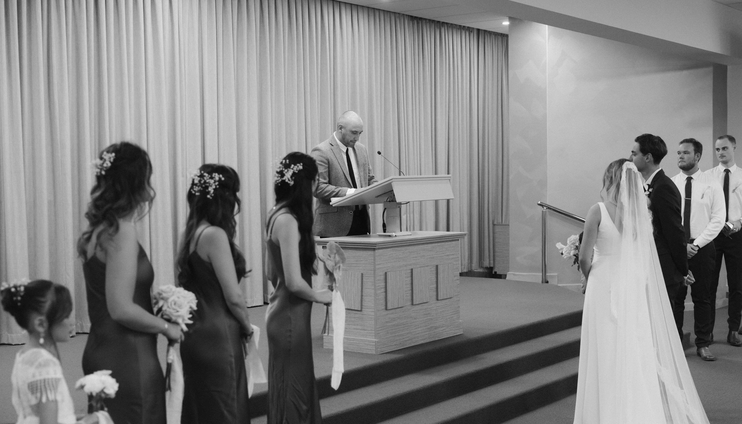 A black and white photo of a wedding ceremony with the bride and groom facing each other. The bride is wearing a white wedding dress and veil, holding a bouquet, and the groom is dressed in a dark suit. Bridesmaids, in matching dresses, stand to the left holding flowers, and groomsmen are lined up to the right, some with hands folded. A man, possibly officiant, stands at a podium, reading or speaking. The setting includes a curtain backdrop and a small set of stairs.