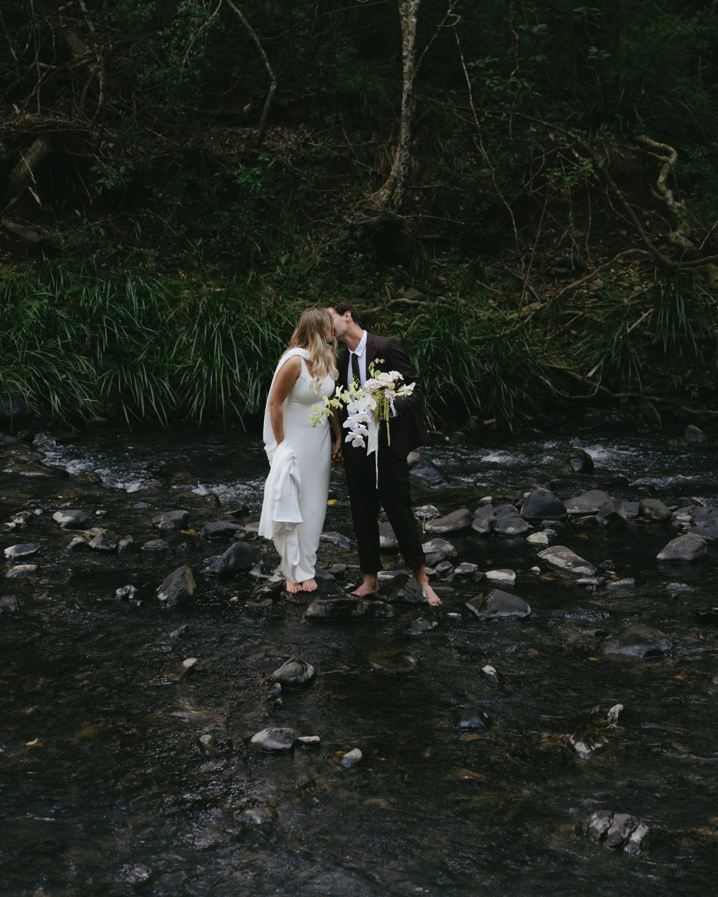 A bride and groom kissing in a river surrounded by greenery.