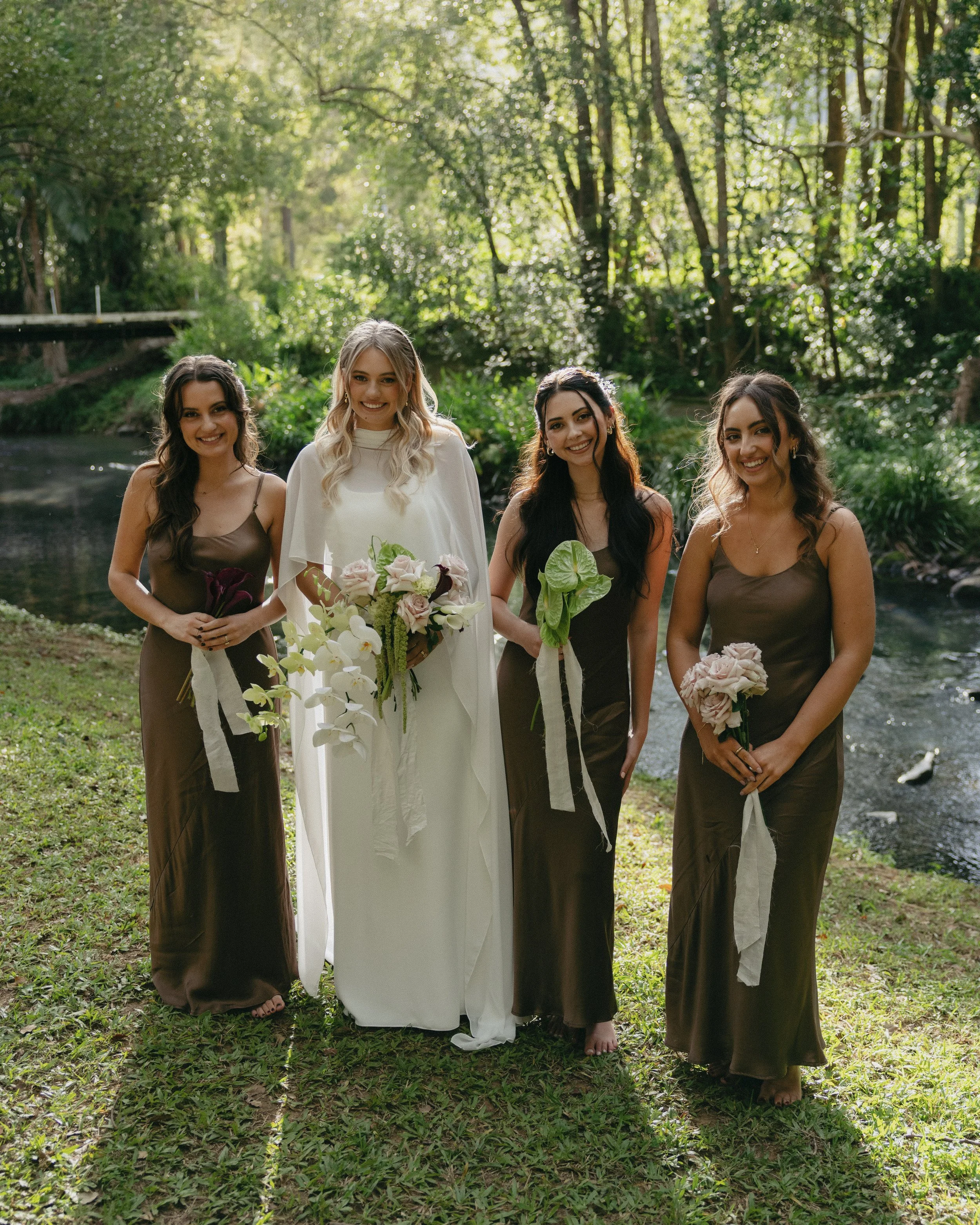 A bride and three bridesmaids standing outdoors near a river with trees in the background, holding bouquets of flowers, dressed in formal attire.
