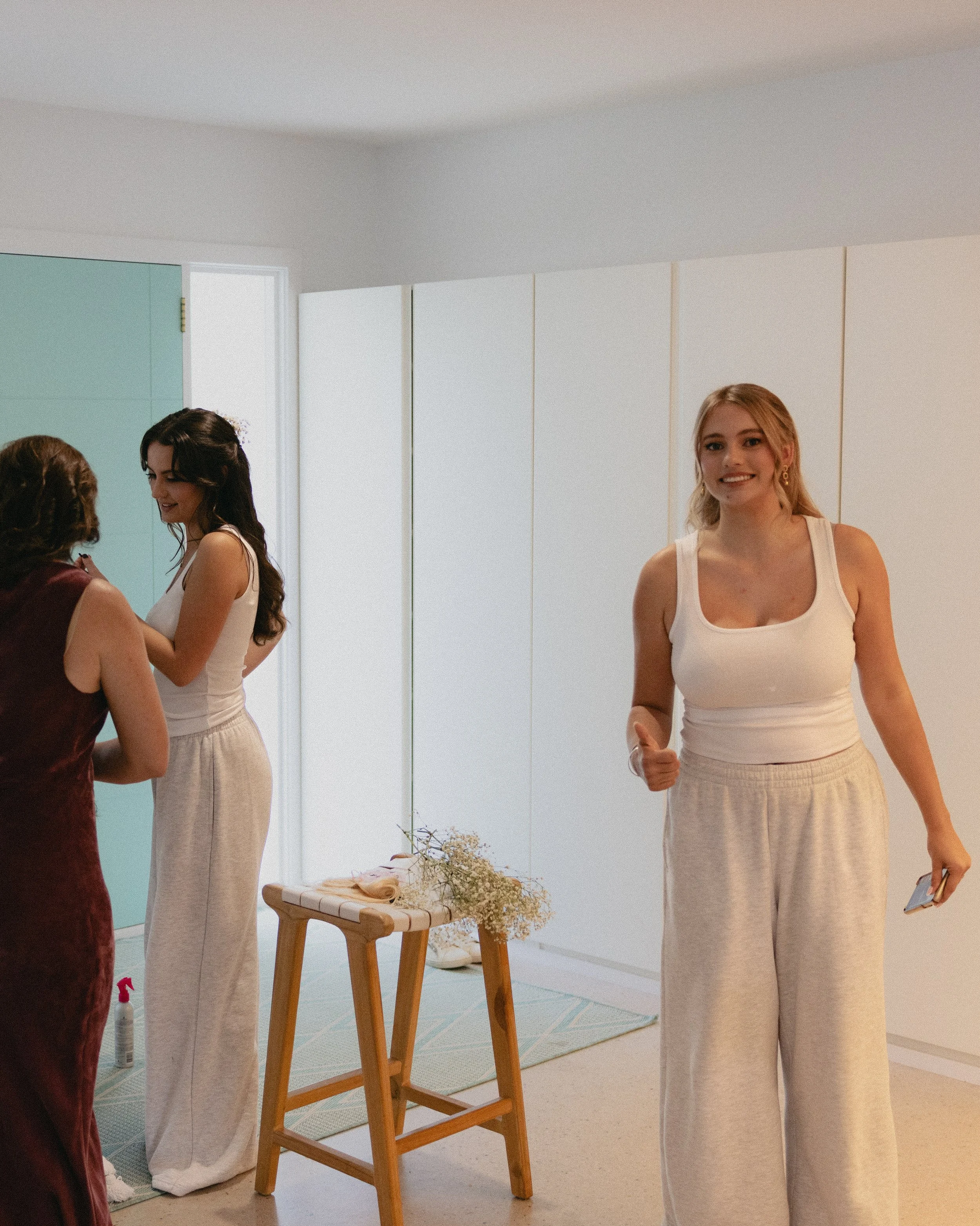 Three women preparing for a wedding in a bright, modern room, with one smiling and holding a phone, and the others adjusting her dress near a small table with flowers and a bottle of sanitizer.