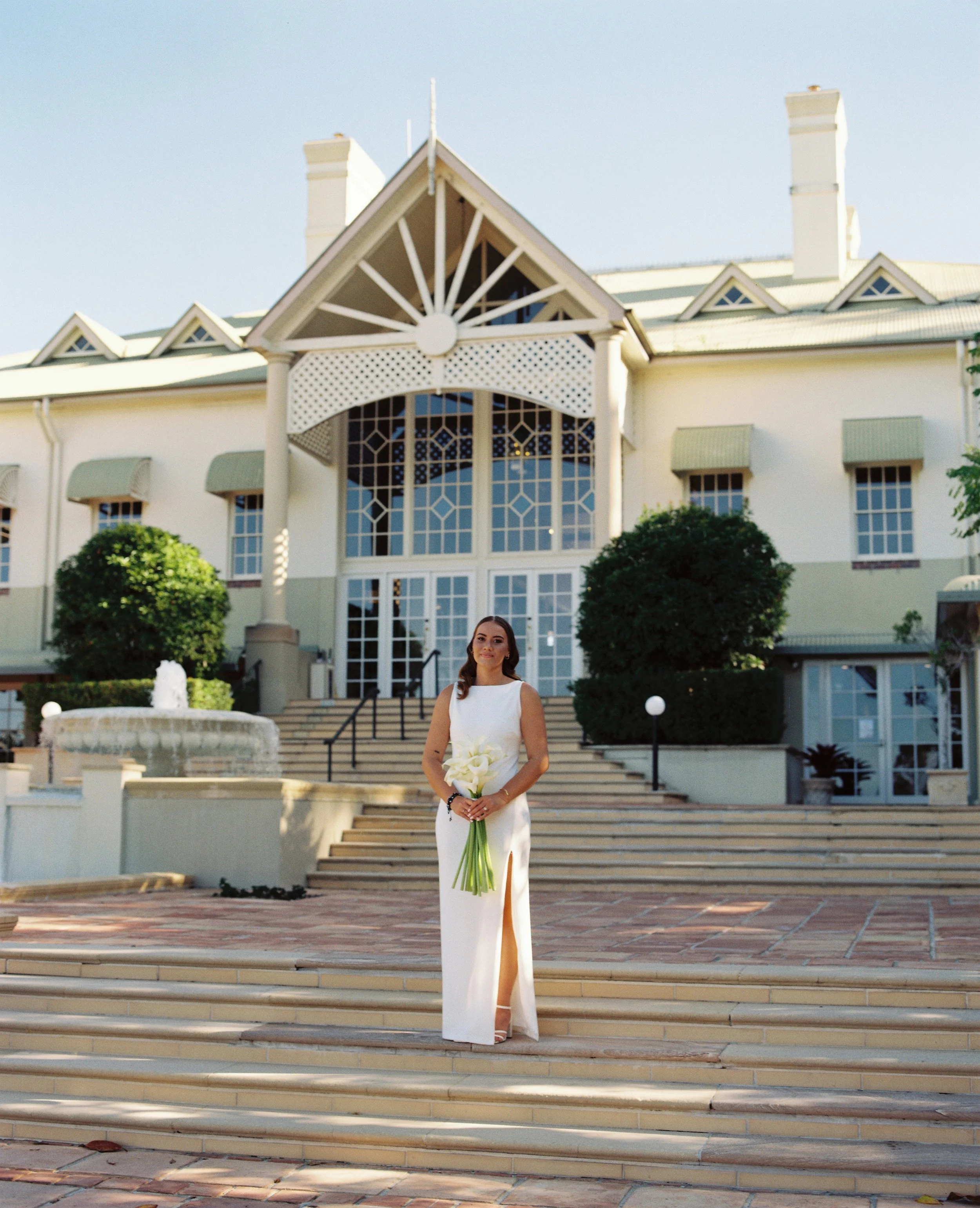 Woman in a white dress holding a bouquet of white calla lilies, standing on steps in front of a large, elegant building with a fountain and landscaped bushes.