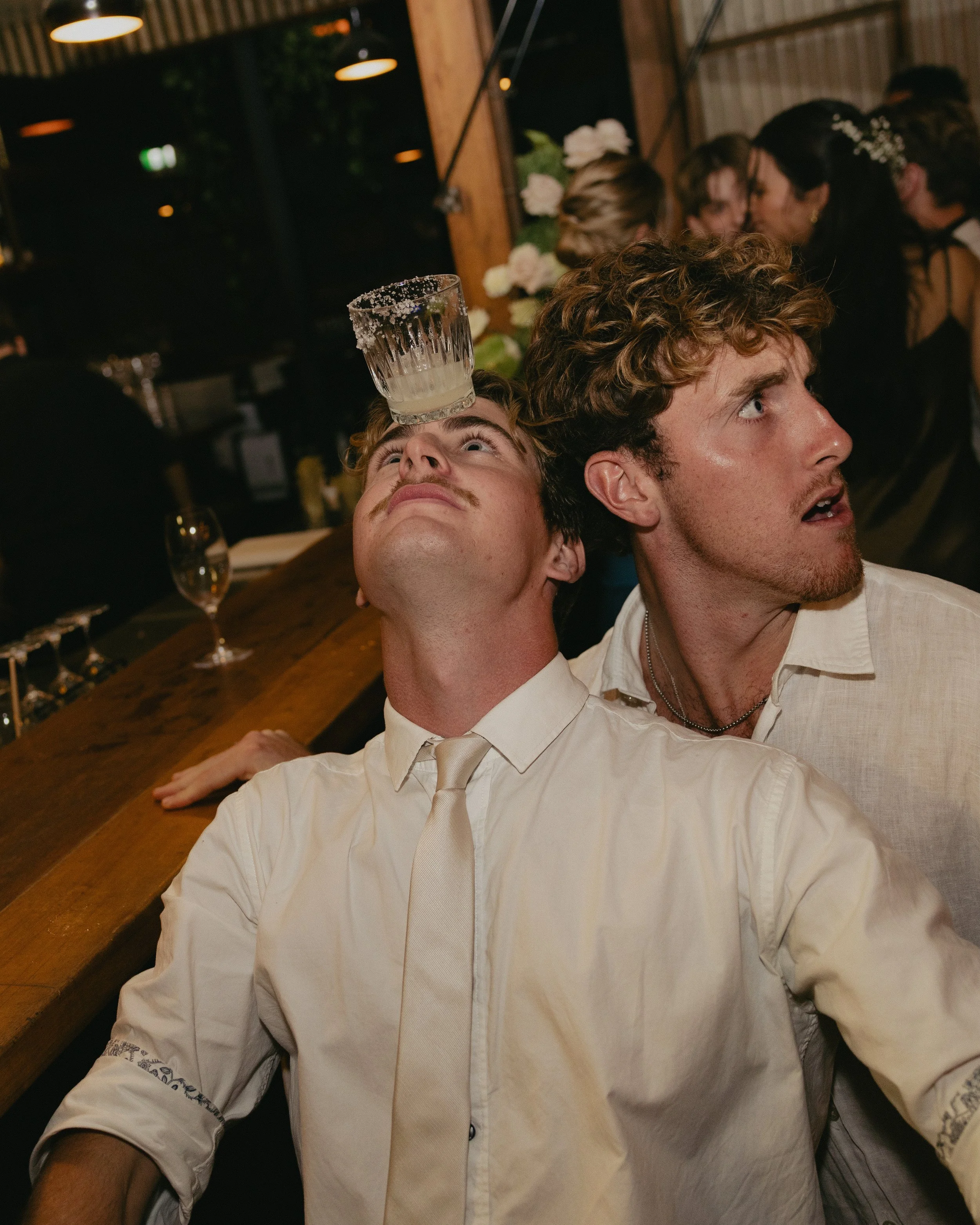Two young men are at a party or celebration, with one man balancing a shot glass on his forehead while looking up, and the other man sitting close beside him, looking to the side. The background shows other people socializing and a wooden wall with flowers.