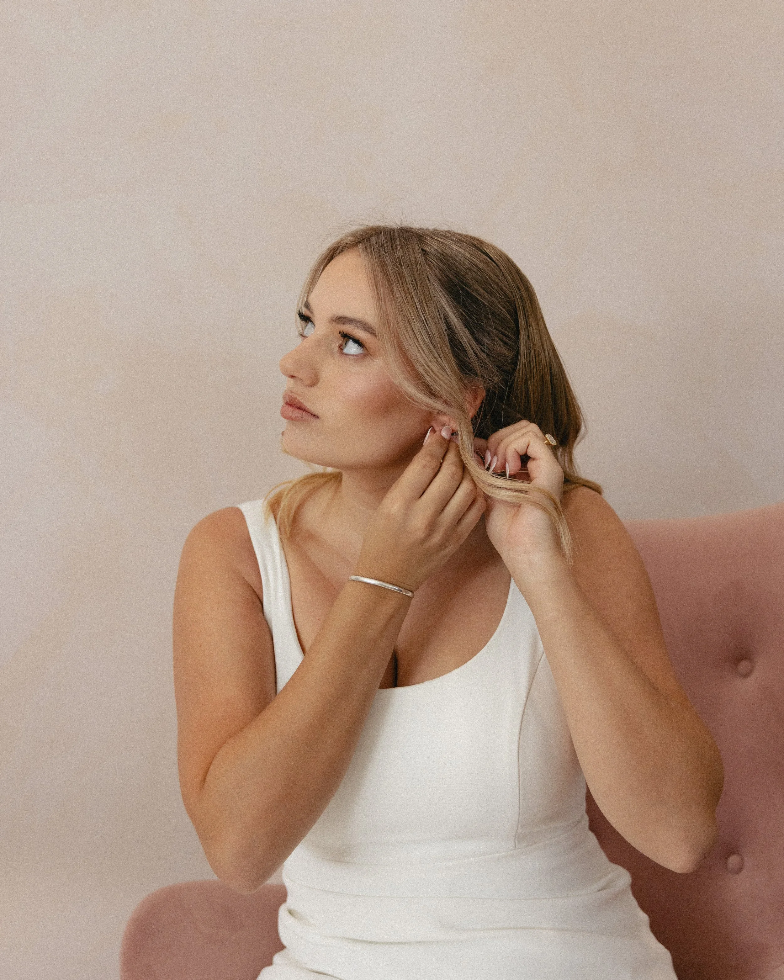 Young woman with blonde hair styling her hair while sitting on a pink upholstered chair, wearing a white sleeveless top, and wearing a bracelet and ring.