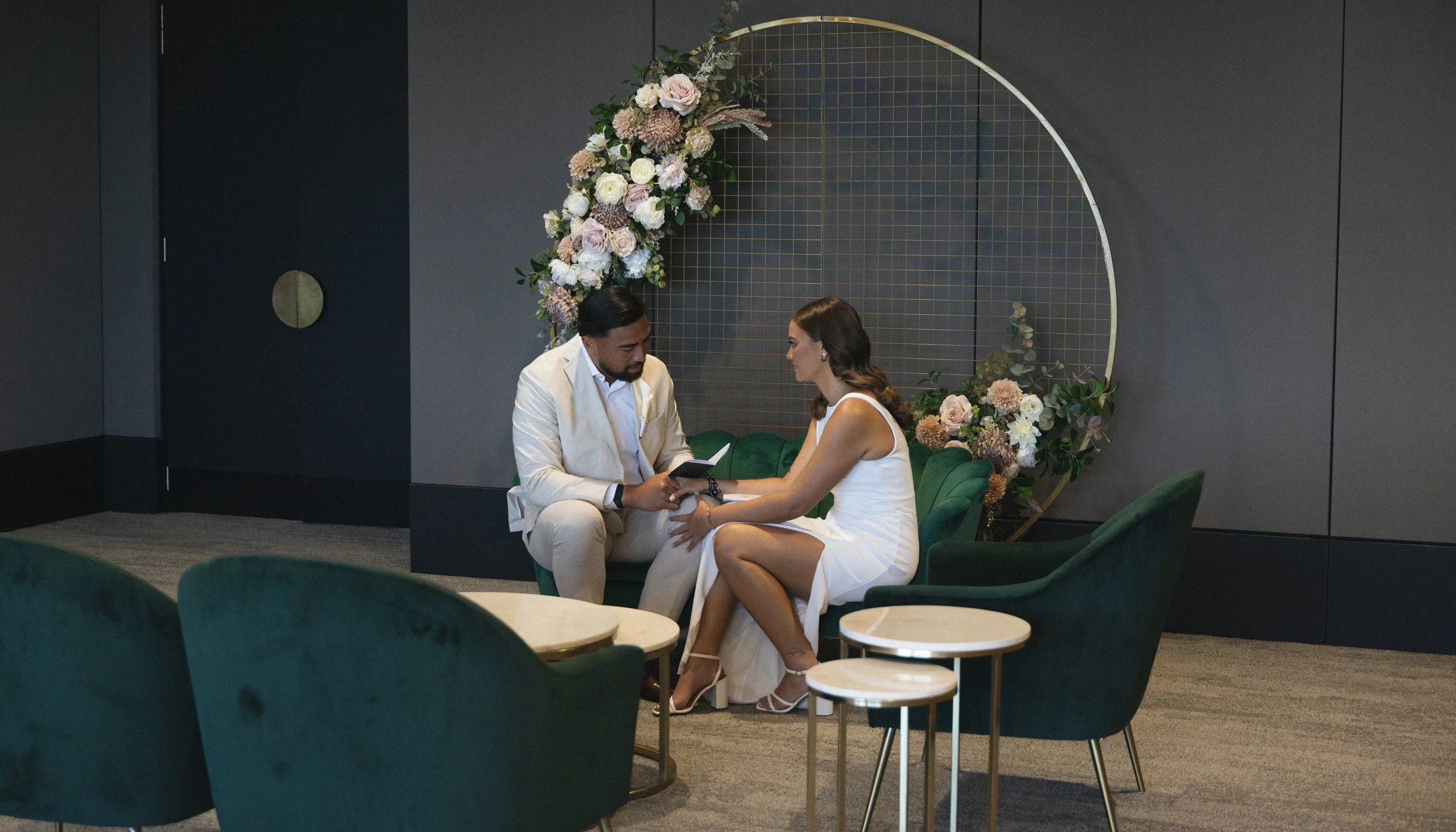 A man and woman sitting on a green couch exchanging rings at a wedding ceremony, with floral decorations and a decorative backdrop behind them.