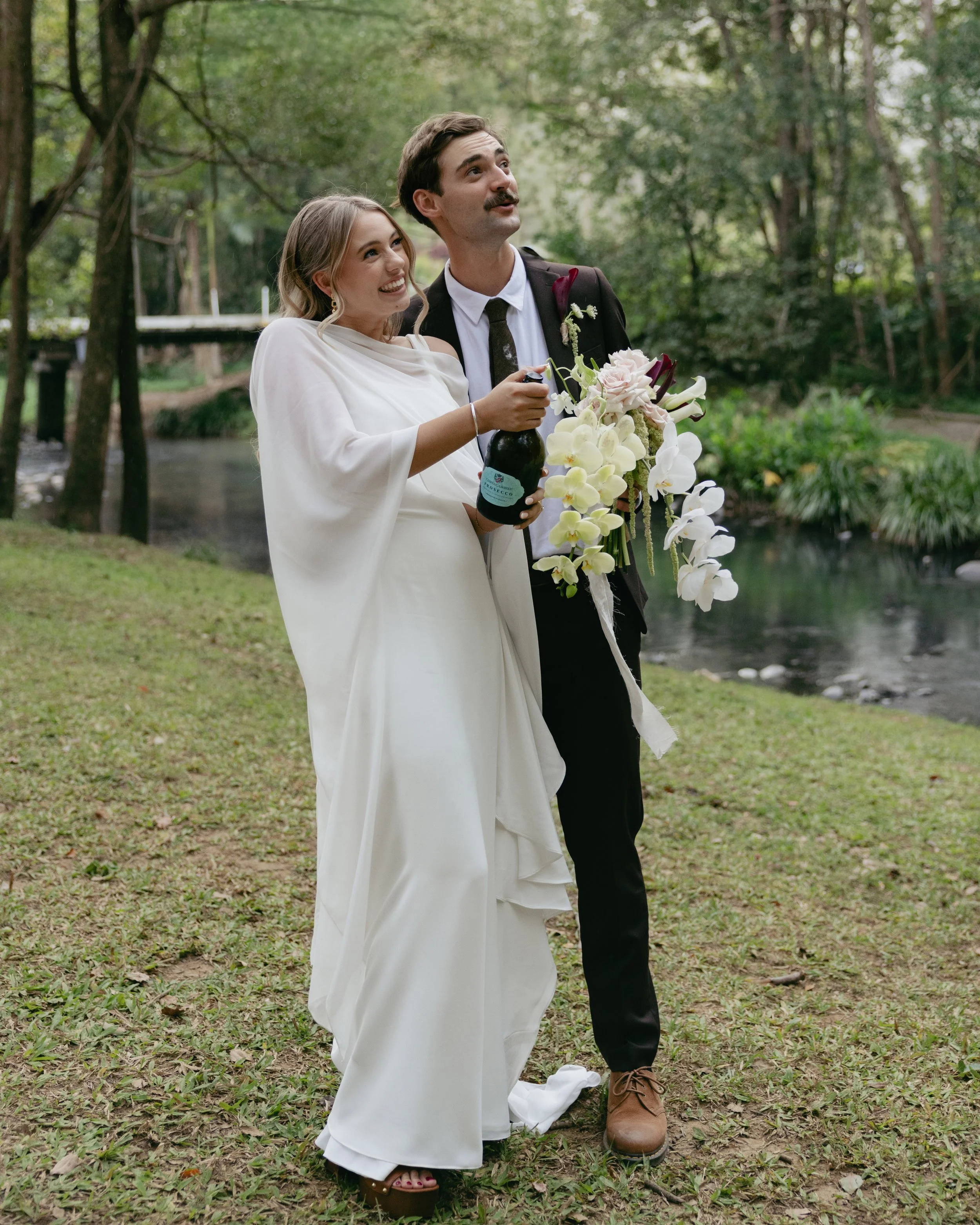 A bride and groom celebrating outdoors near a river, with the bride holding a bottle of champagne and wearing a white wedding dress, and the groom holding a bouquet of flowers, both smiling and enjoying the moment.