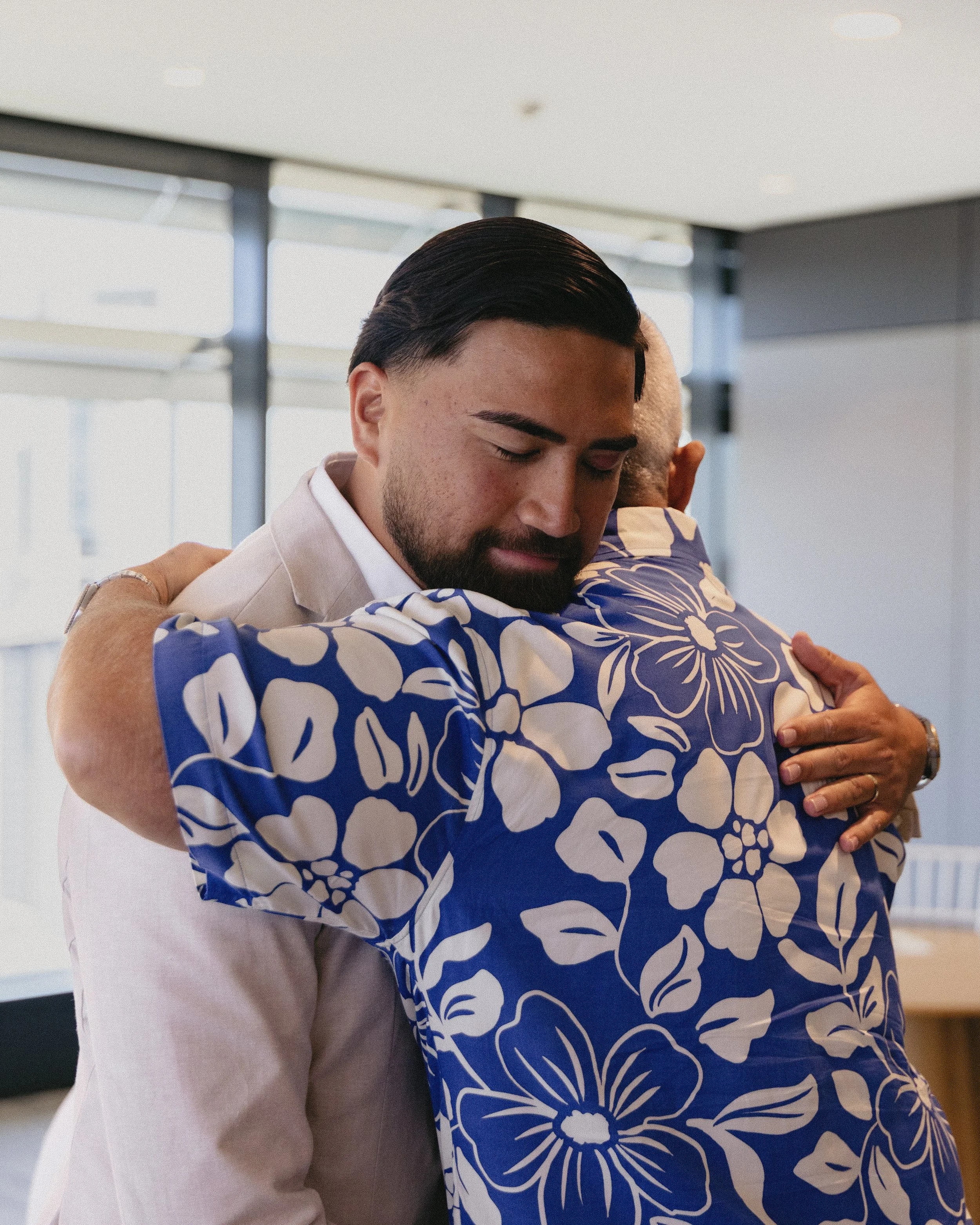 Two men hugging warmly in an indoor setting, with one wearing a white suit and the other in a blue Hawaiian shirt with white floral patterns.