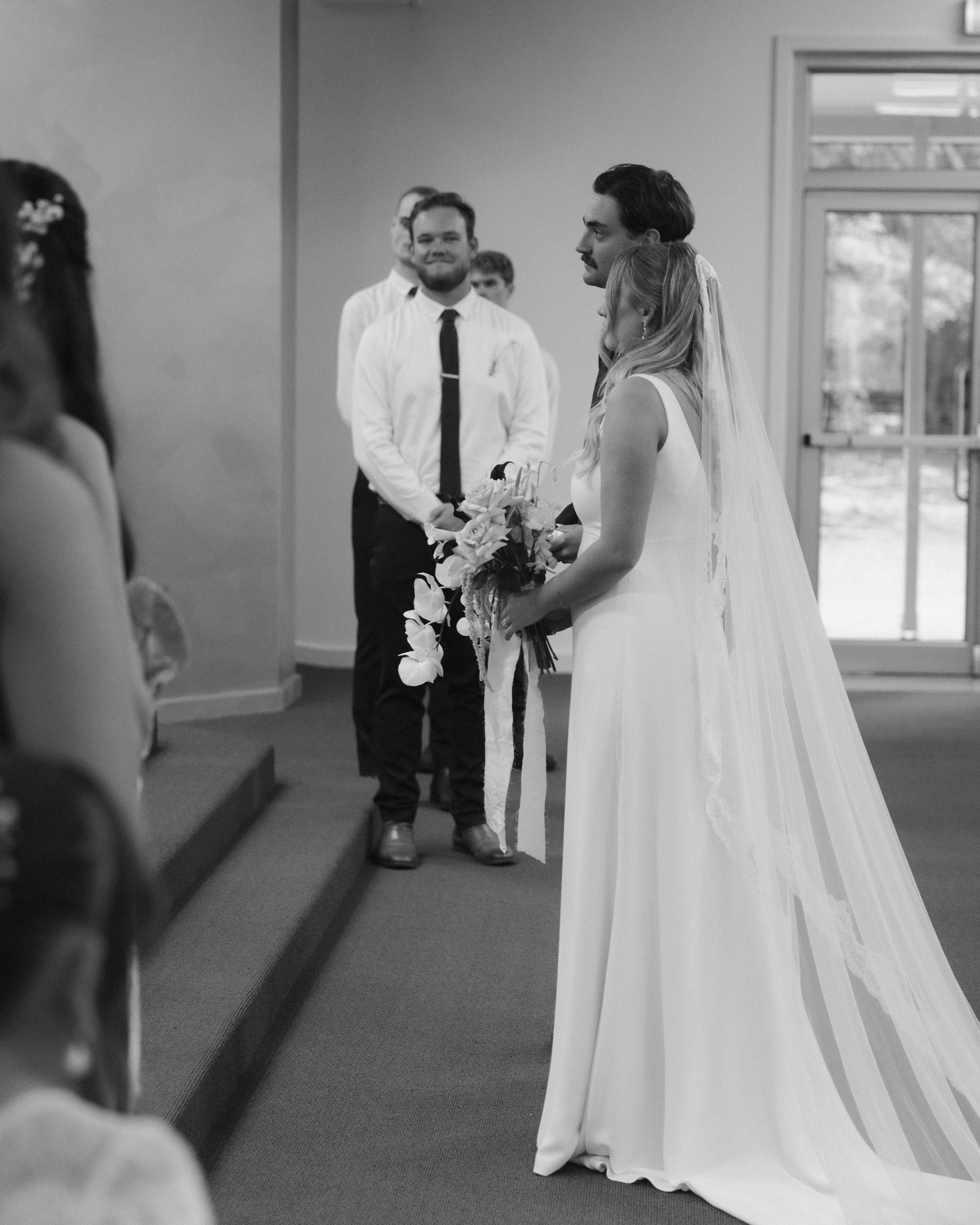 Bride in a white wedding dress holding a bouquet, standing at the altar during a wedding ceremony, with the groom and guests in the background.