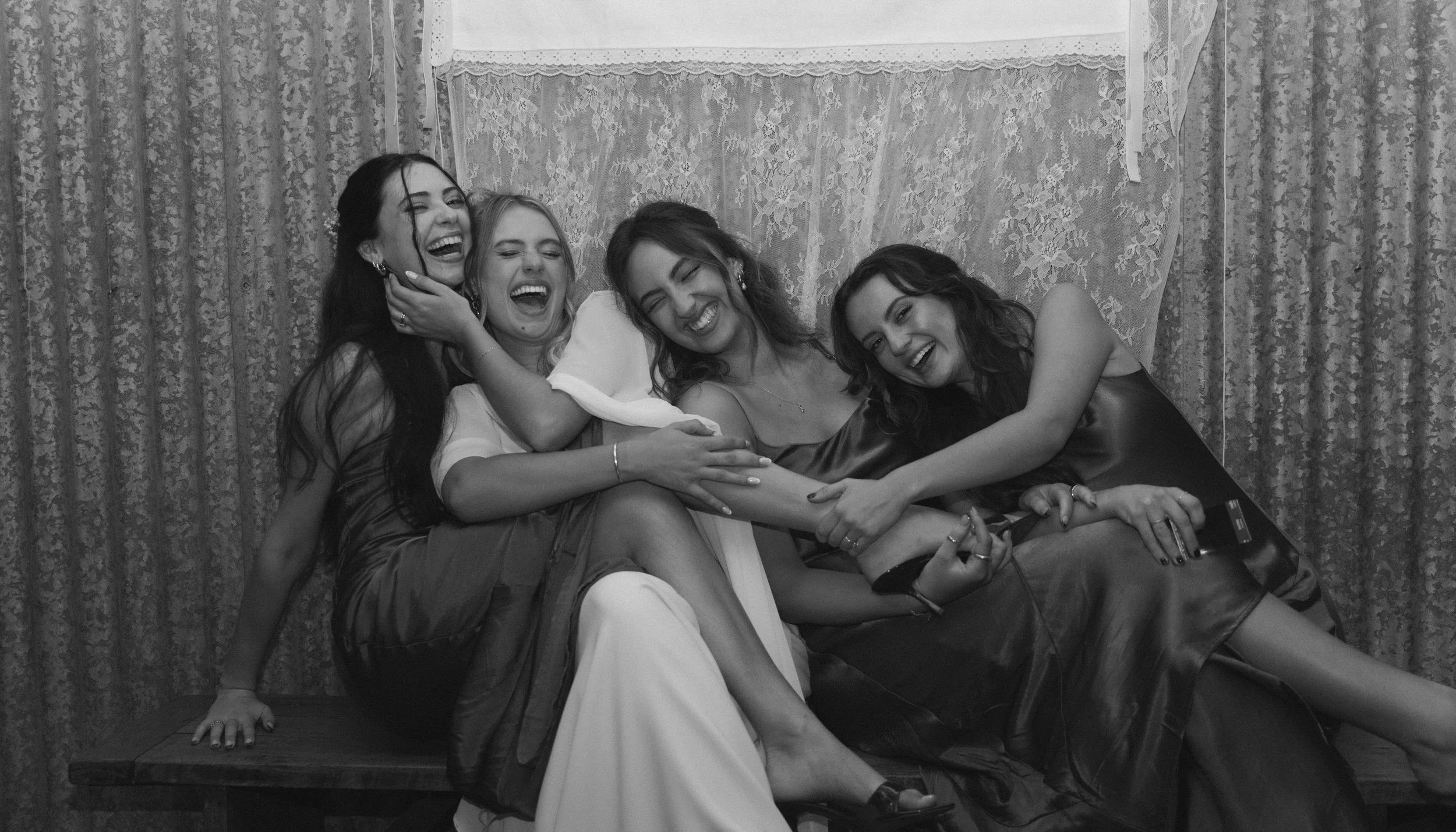 Five women sitting on a wooden bench, laughing and hugging each other, in a room with lace curtains and patterned drapes.