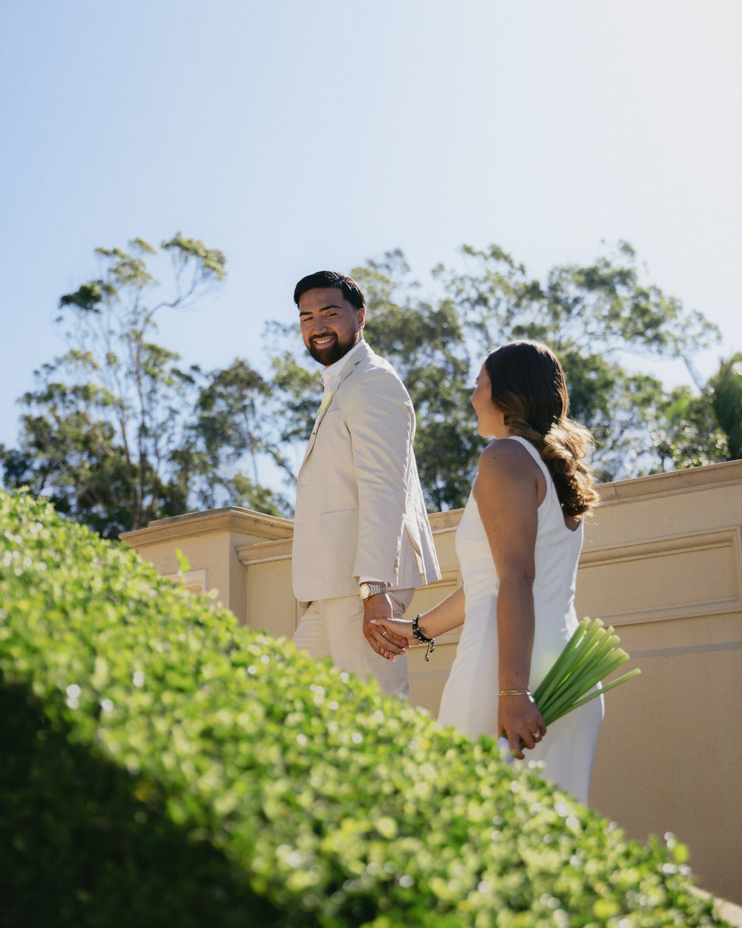 A couple holding hands, dressed in white wedding attire, standing outdoors on a sunny day with trees in the background. The woman is holding a bouquet of green flowers.