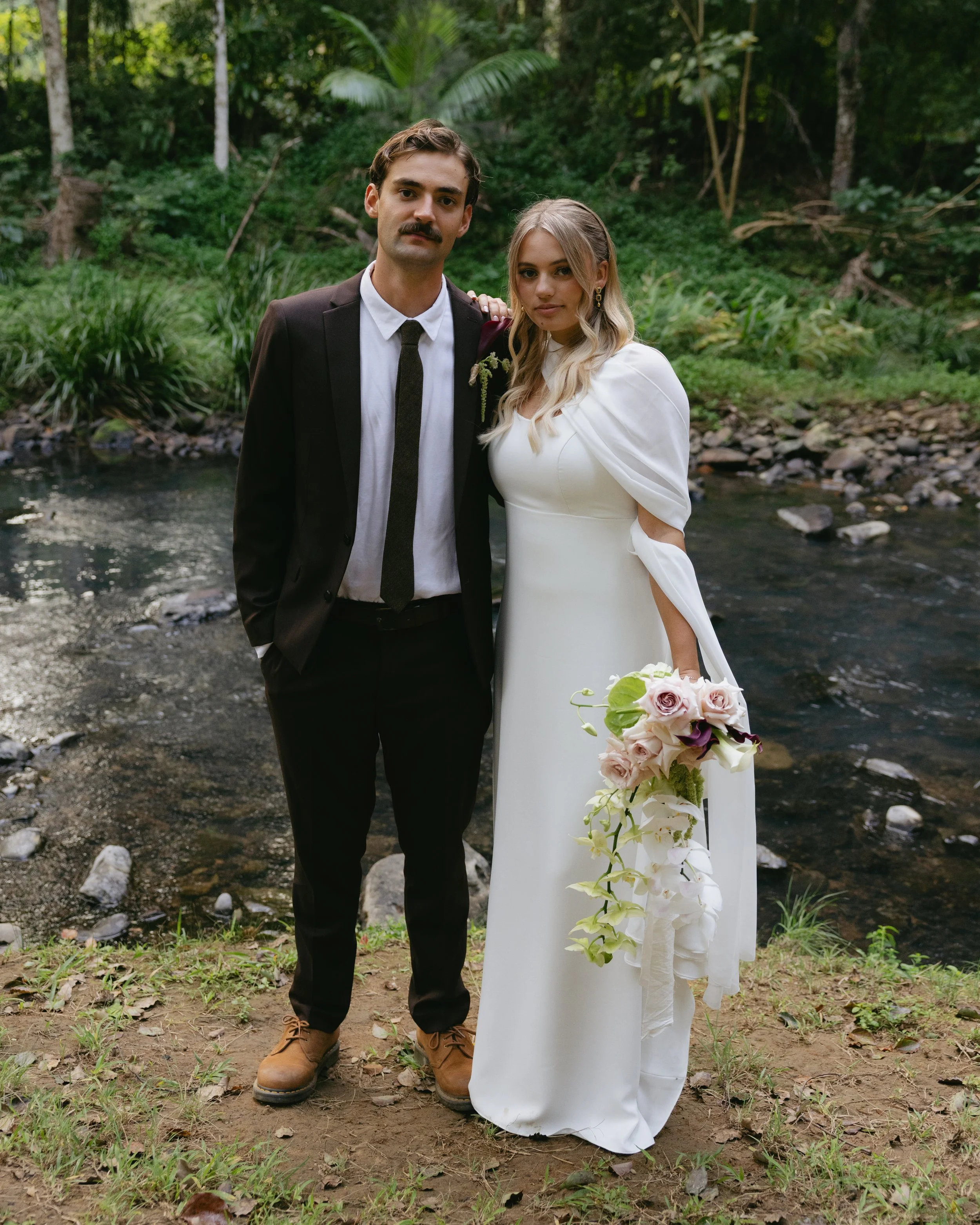 A man and a woman in wedding attire standing by a creek in a lush, green forest. The woman is holding a bouquet of flowers.