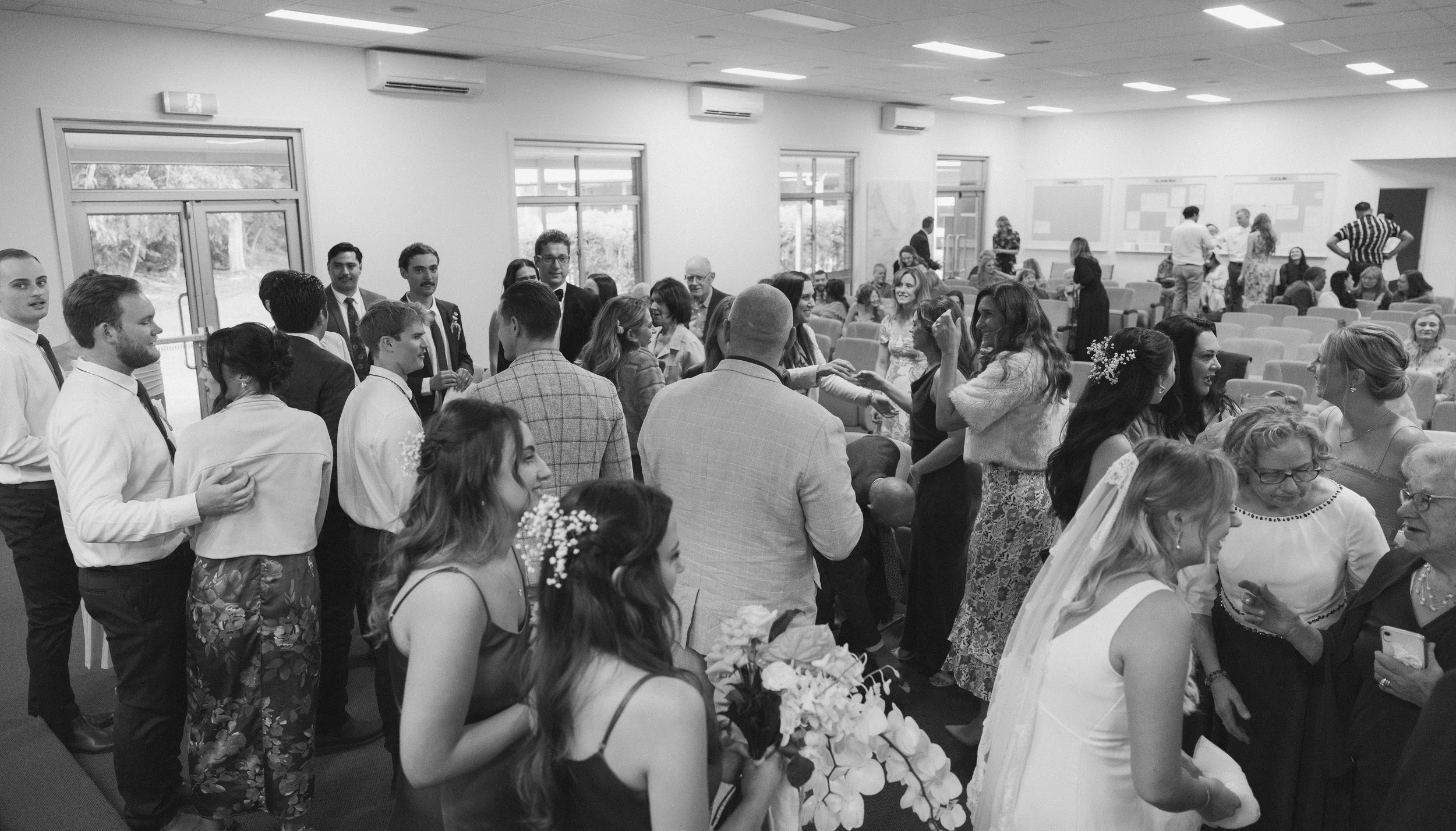 A black and white photo of a wedding reception with guests socializing, dancing, and a bride holding a bouquet in a decorated indoor venue.