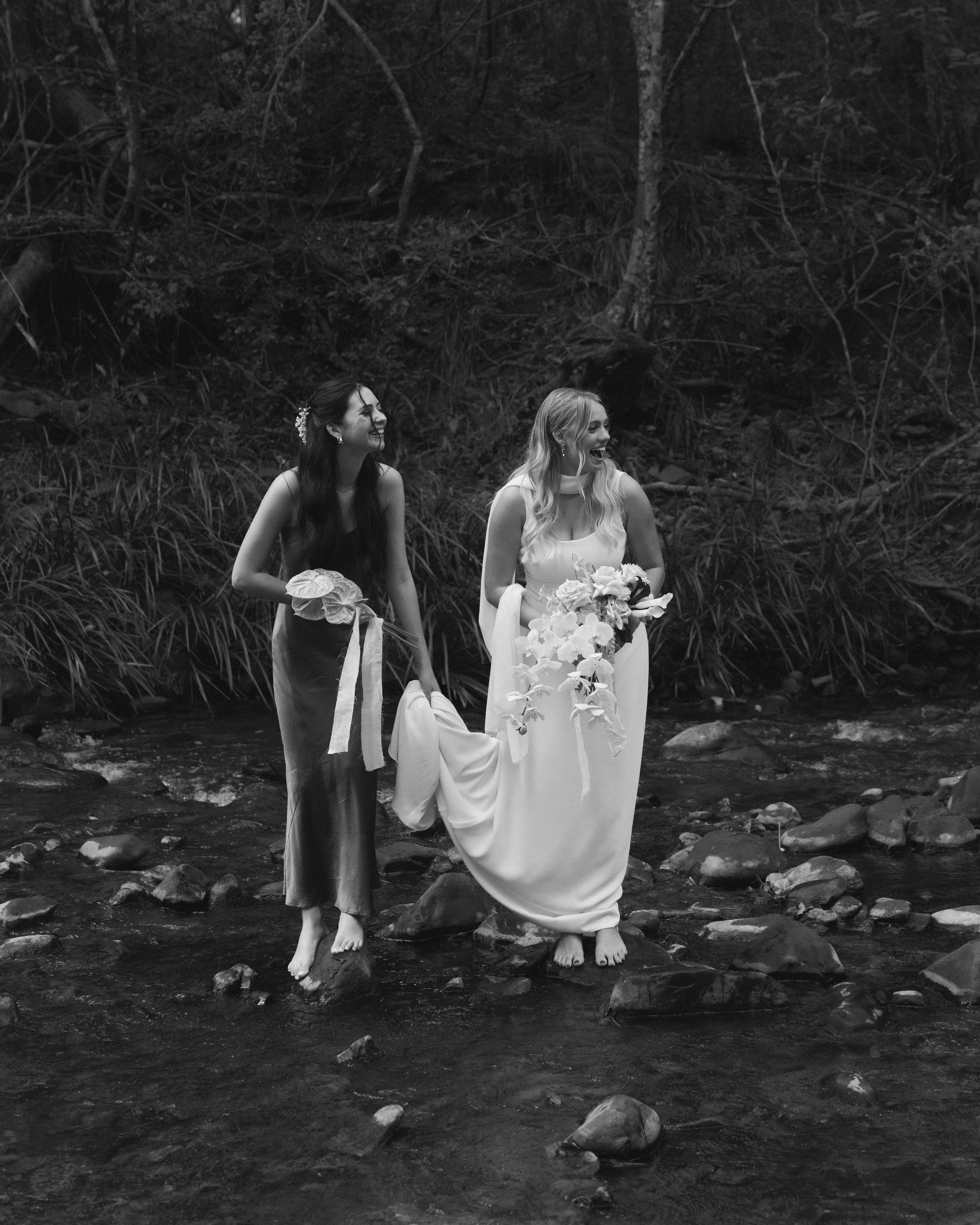 Two women in wedding dresses standing barefoot in a creek, one holding a bouquet of flowers and the other holding a ribbon, smiling and looking at each other, surrounded by trees and nature.